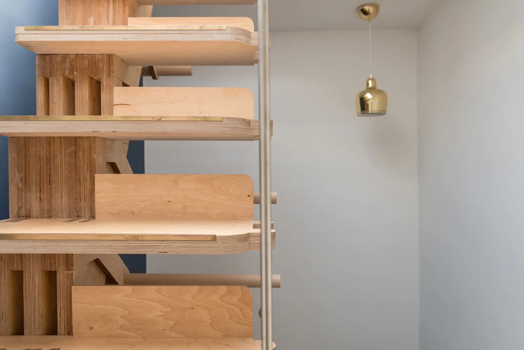 A custom plywood staircase features brass metal strip inlays on the tread edges with a gold pendant light hanging in the background.
