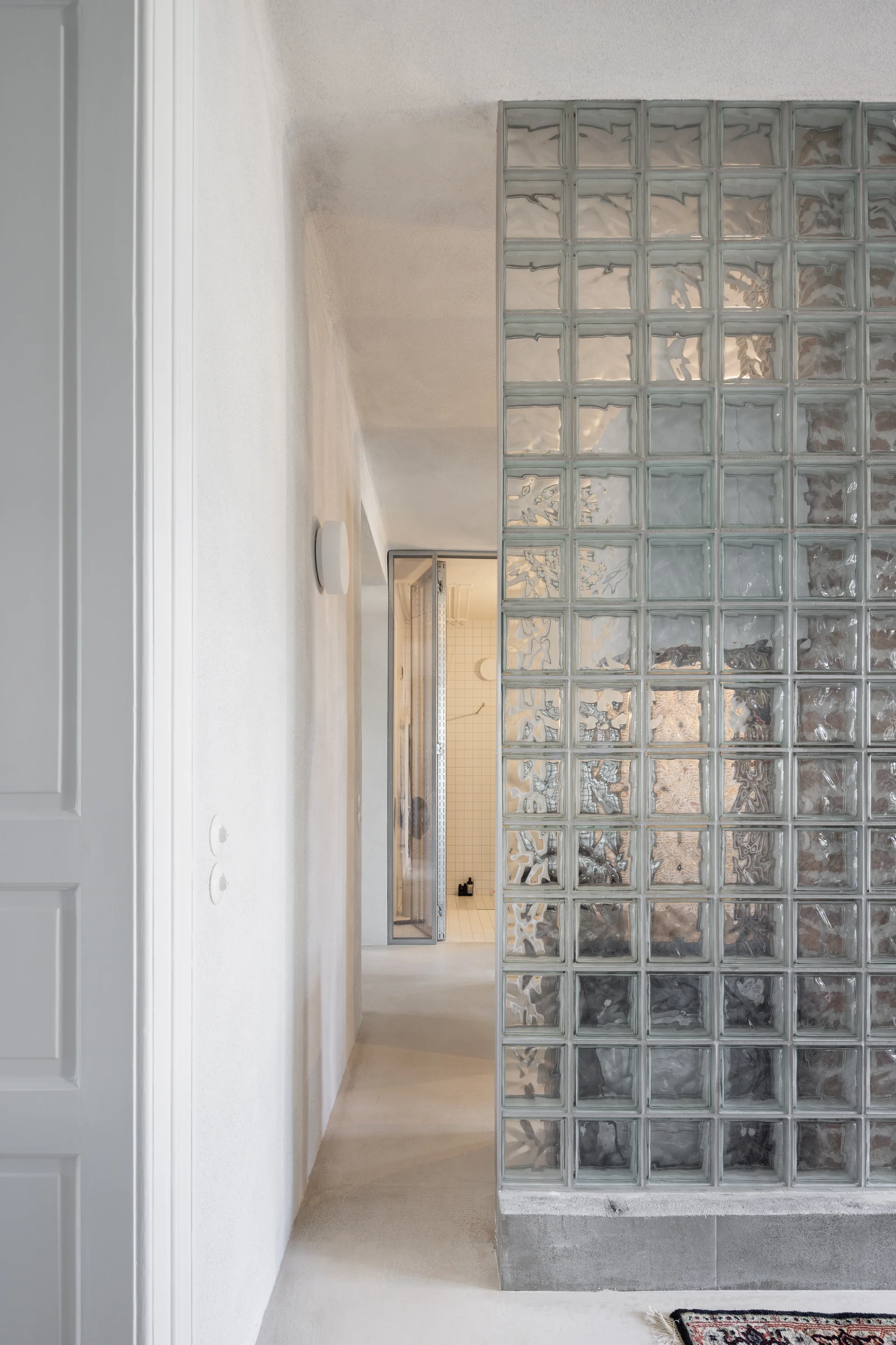 A minimalist white hallway featuring a translucent glass block partition wall on a concrete base next to a plastered corridor.