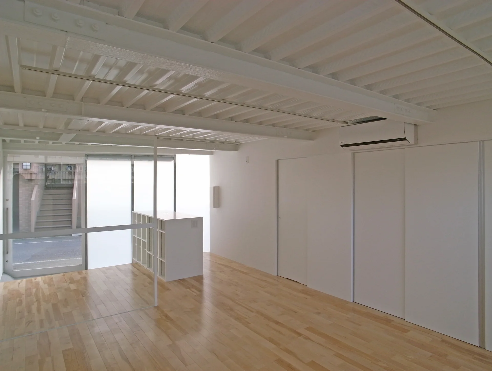 A minimalist room with light wood flooring, white walls, sliding doors, and an exposed white steel deck ceiling.