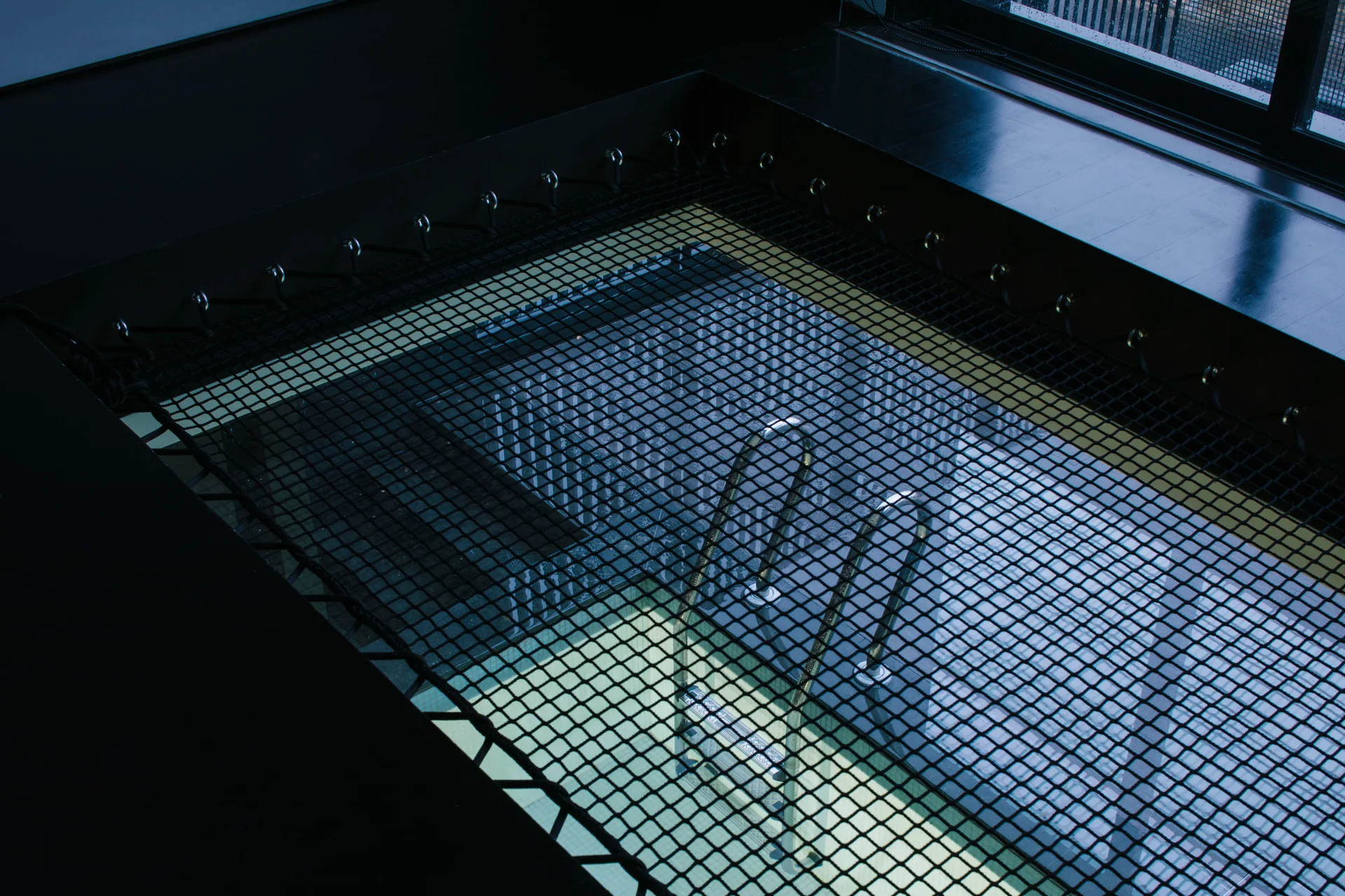 An overhead view of an indoor swimming pool covered by a tight black safety net with a metal pool ladder visible in the water.