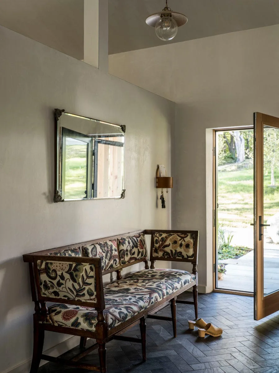 A rustic entryway features an upholstered floral bench, a rectangular mirror, and a dark herringbone slate floor next to an open glass door.