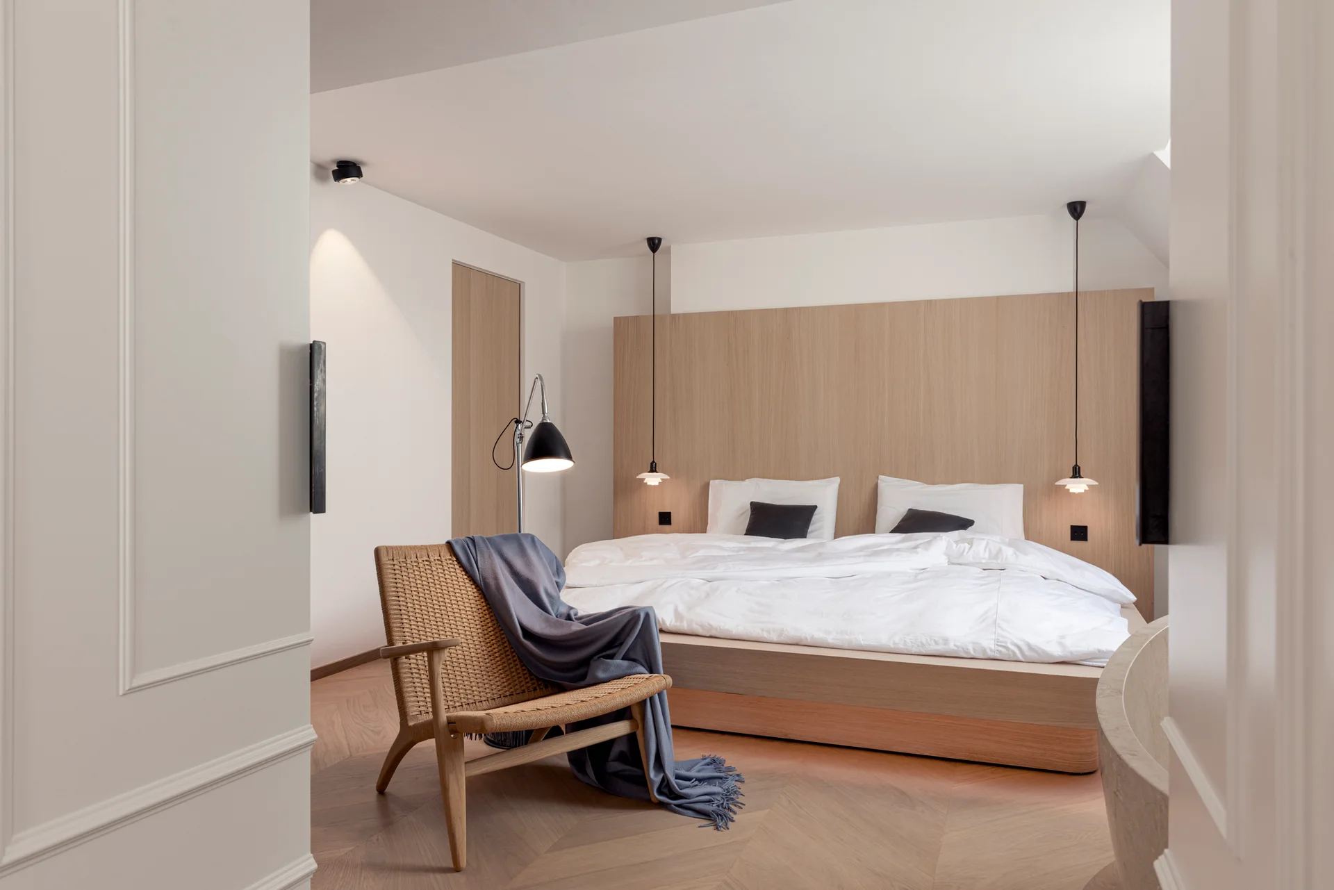 A minimalist bedroom featuring a large light wood wall panel headboard, a low bed, chevron wood flooring, and a woven armchair.
