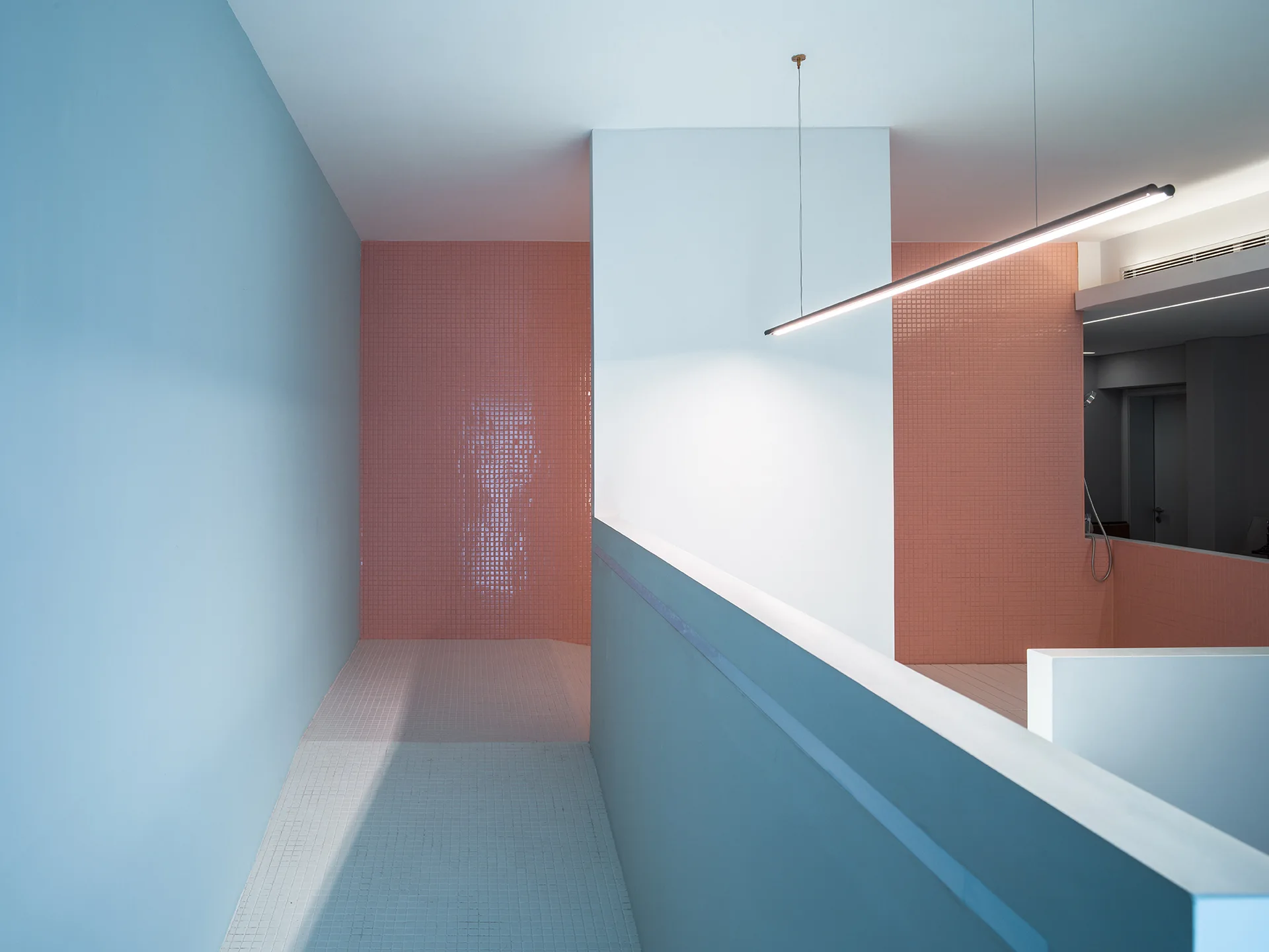 A minimalist hallway features a pastel blue wall, a peach-colored square mosaic tile wall, and a slim linear pendant light fixture.