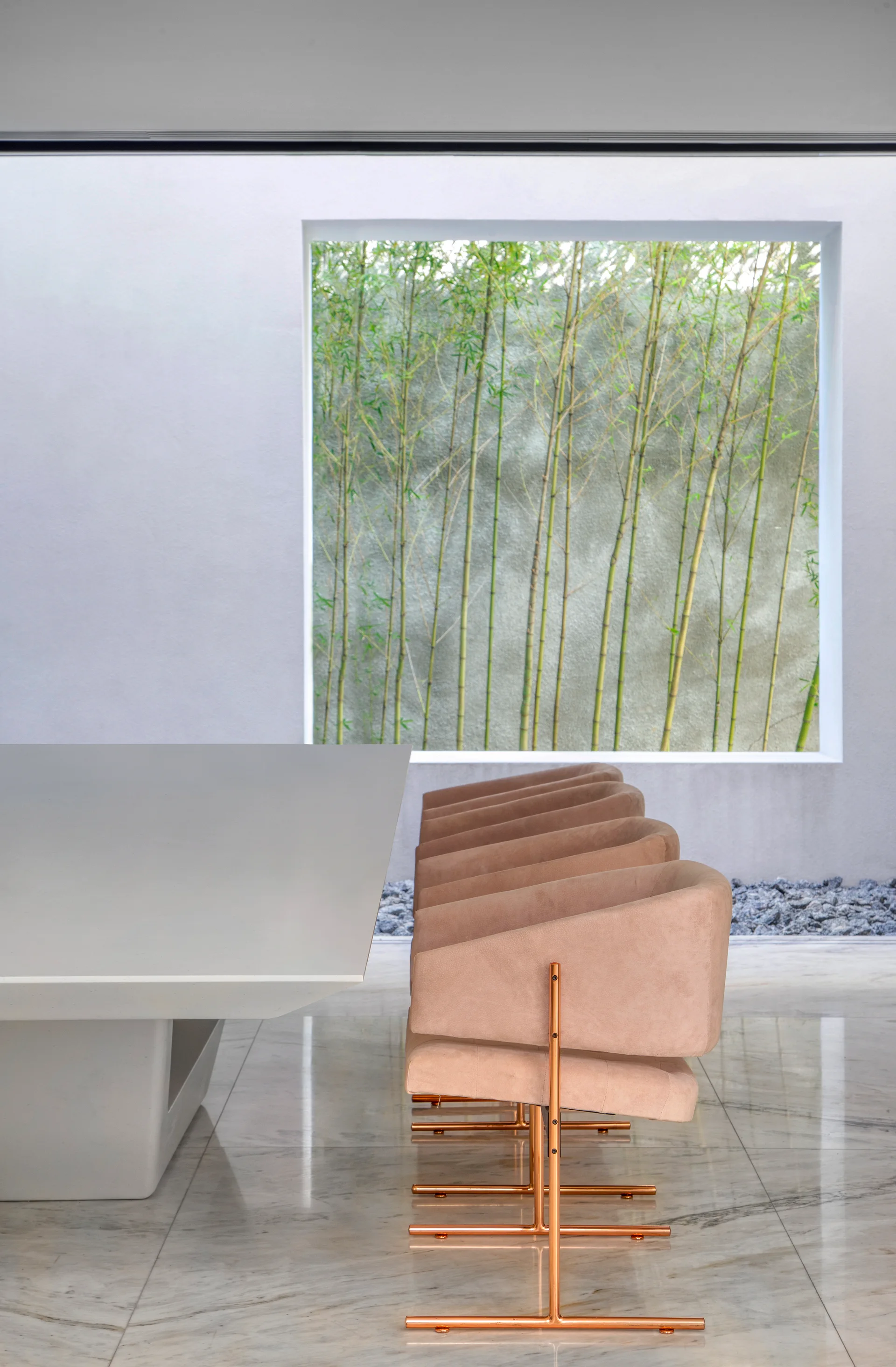 A minimalist dining area features a white stone table, blush pink chairs with copper frames, and a large window looking out at green bamboo.
