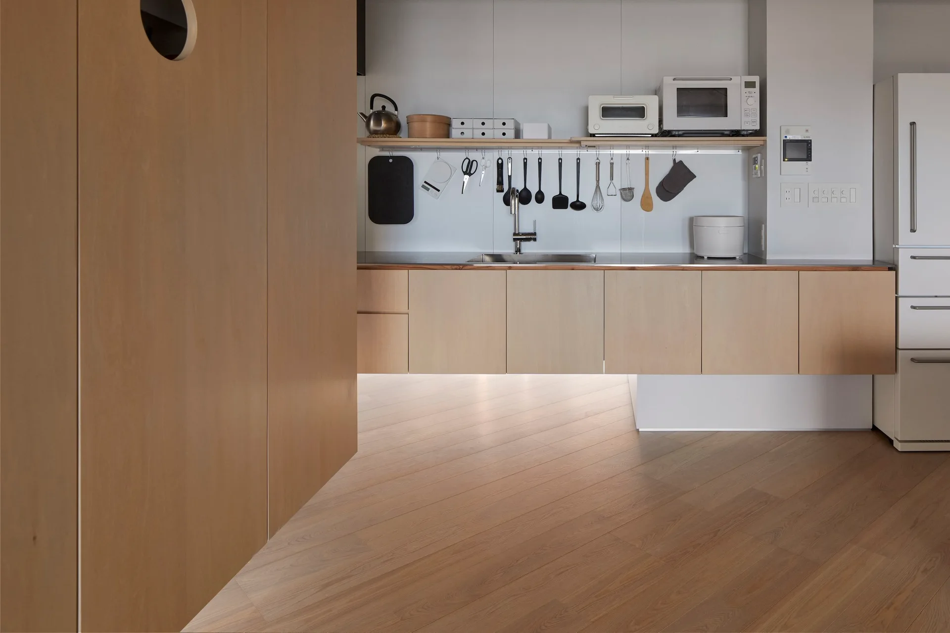 A minimalist kitchen features floating light wood lower cabinets, a stainless steel sink, and an open wooden shelf with hanging utensils.
