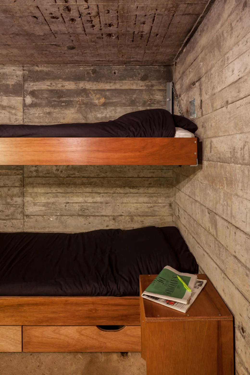 A two-tier wooden bunk bed with black linens is built against textured concrete walls next to a matching bedside table.