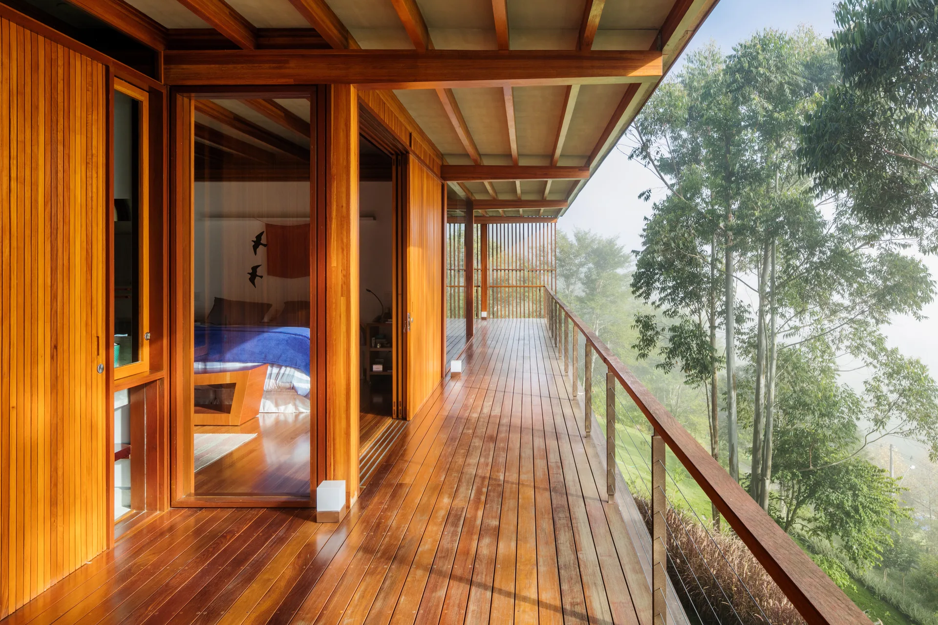 A spacious wooden balcony features polished plank flooring, vertical slat siding, a metal cable railing, and sliding glass doors opening to a bedroom.