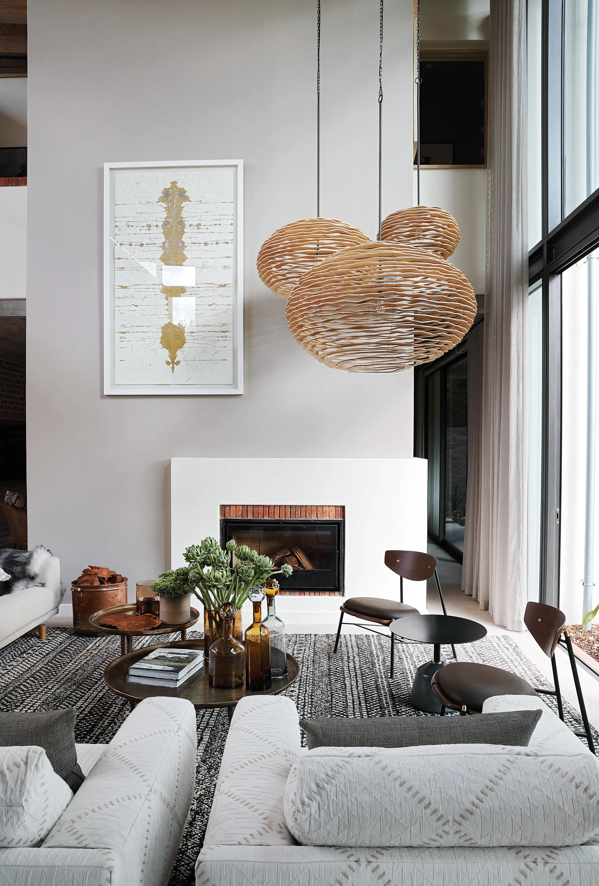 A contemporary living room features textured white sofas, a white brick fireplace, and three large woven wood pendant lights suspended from a high ceiling.