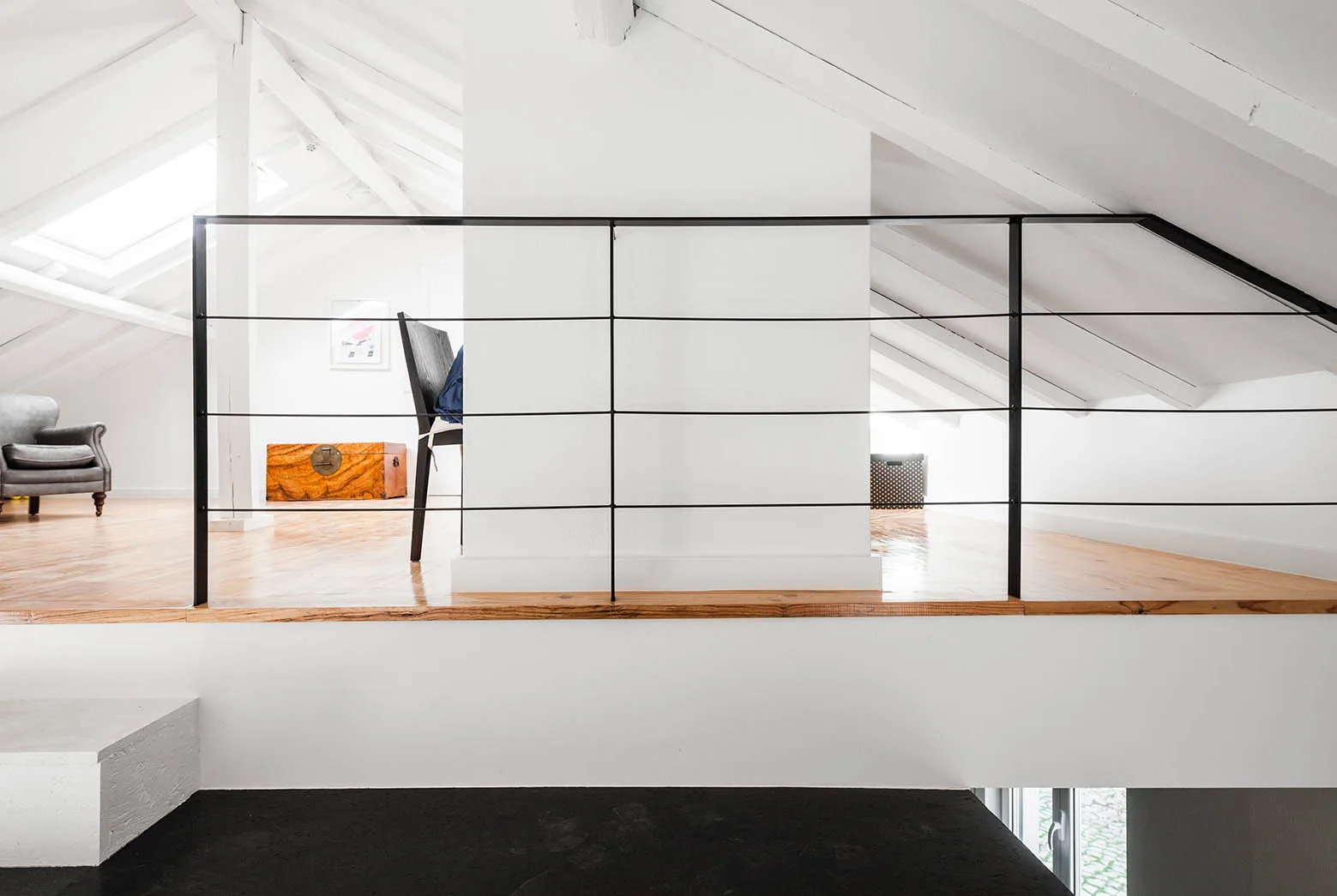 An attic mezzanine features a black horizontal metal railing, warm wood floors, and a white sloped ceiling with exposed painted beams.