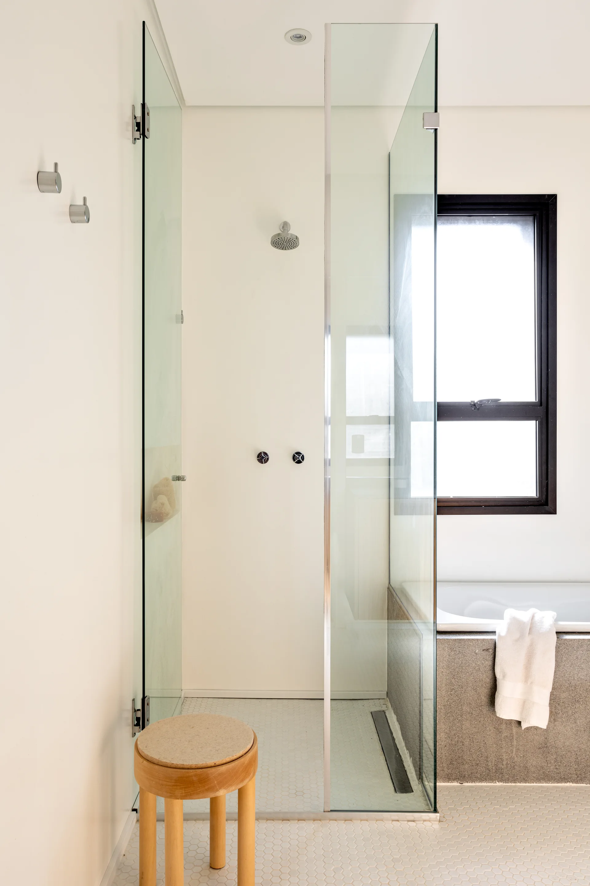 A minimalist bathroom featuring a frameless glass shower enclosure, a built-in tub with a grey stone-look surround, and white hexagon tile flooring.