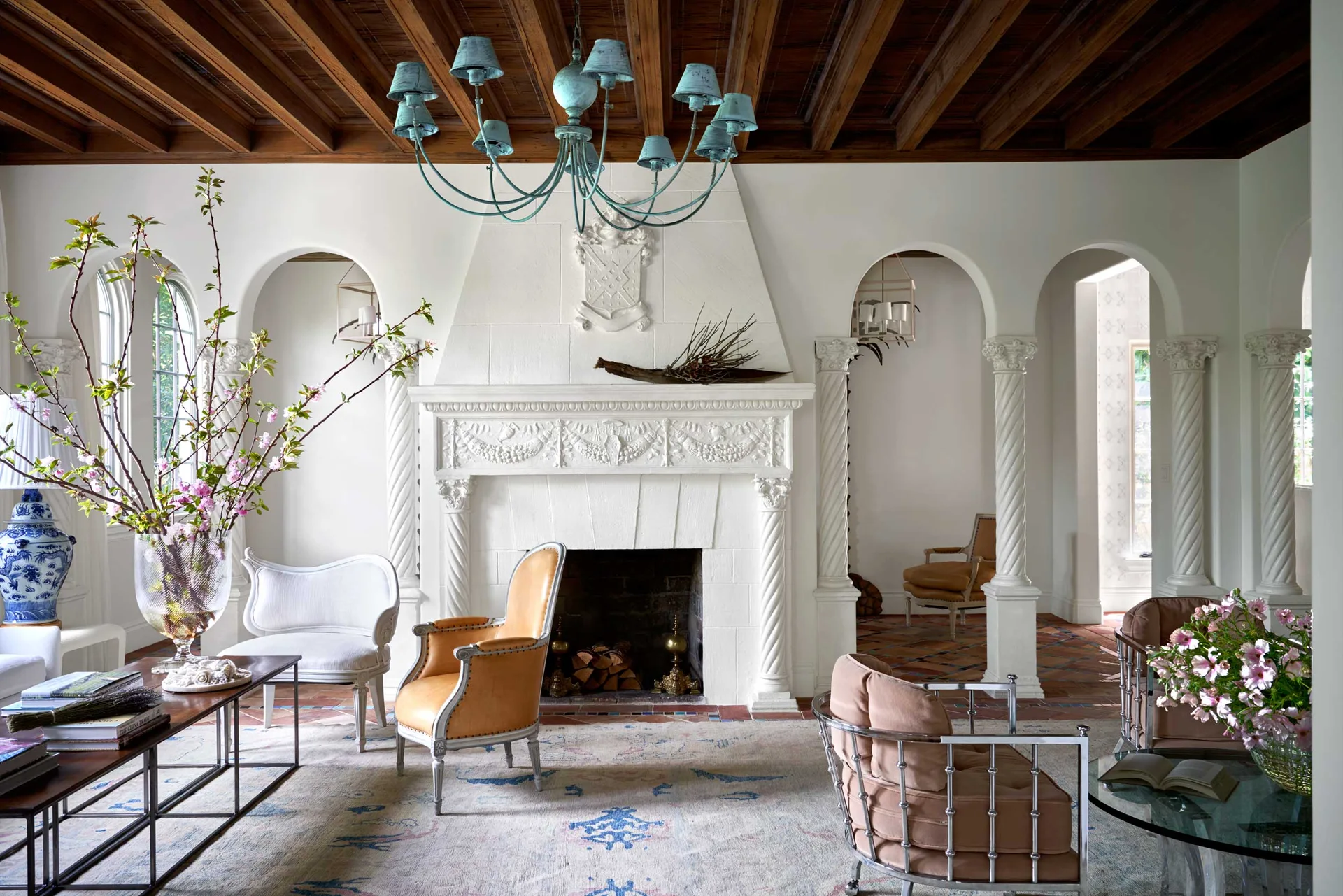 A Mediterranean living room features an ornate carved white fireplace flanked by arched doorways with spiral columns under a dark wood slat ceiling.