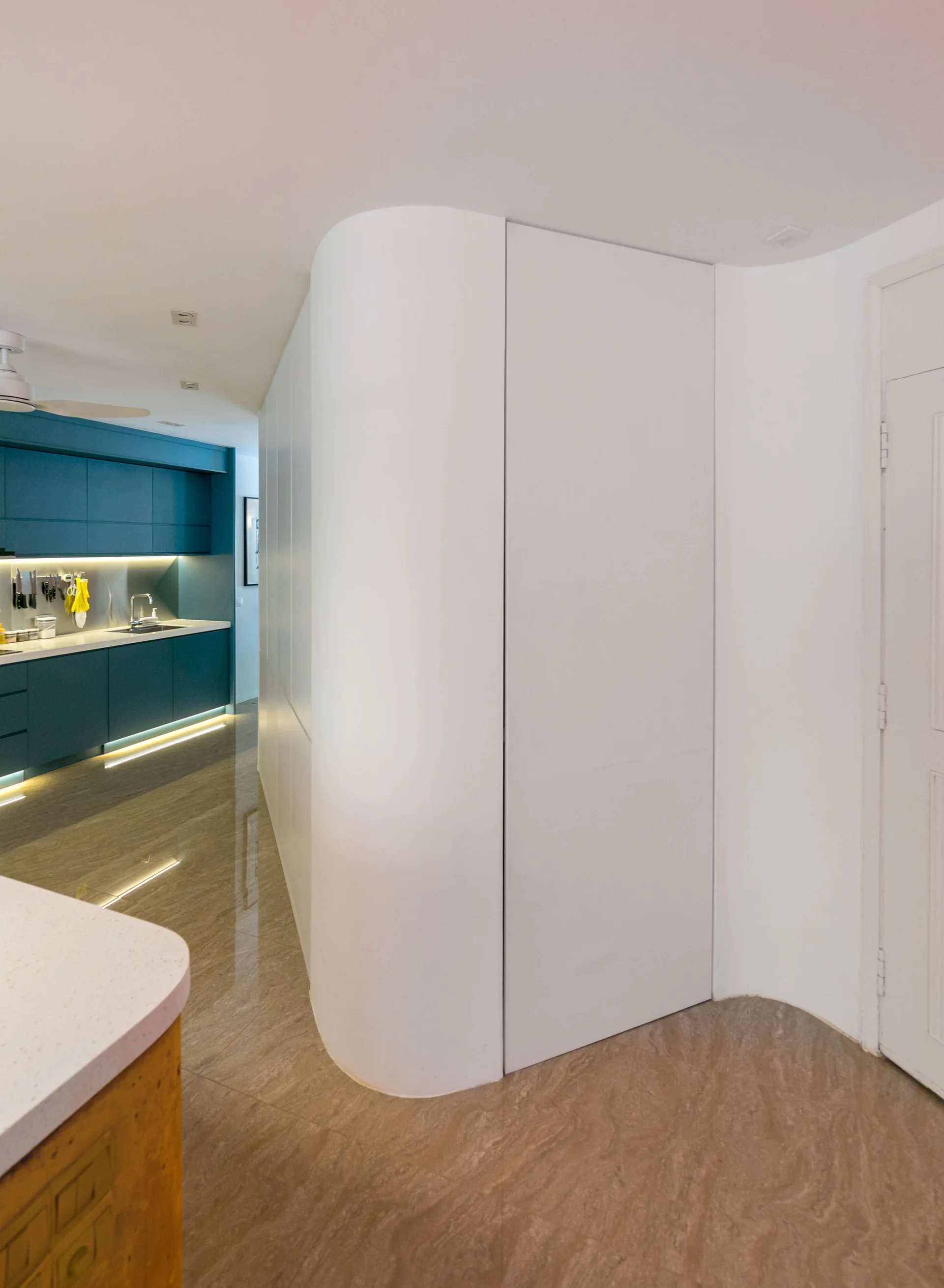 A modern kitchen with deep teal cabinetry, under-cabinet lighting, a curved white partition wall, and glossy beige stone flooring.