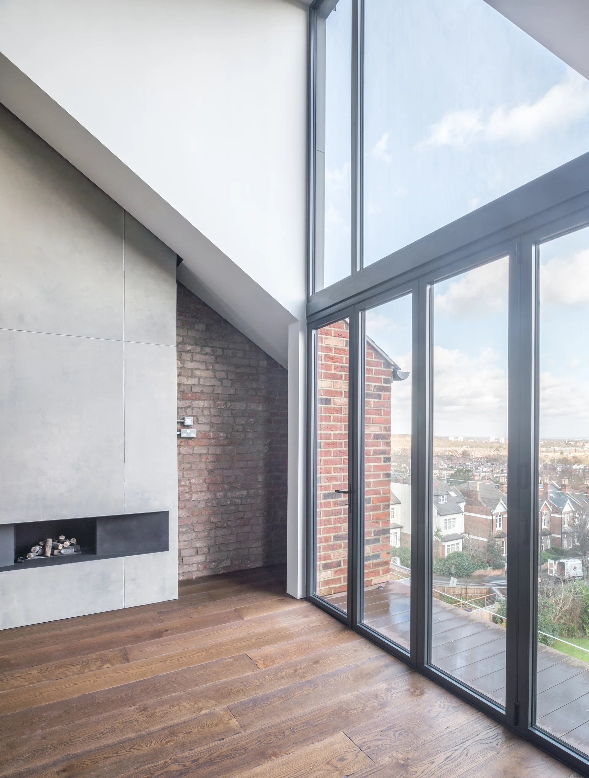 A minimalist living room features a grey concrete fireplace, an exposed brick wall, wood plank flooring, and large floor-to-ceiling windows.