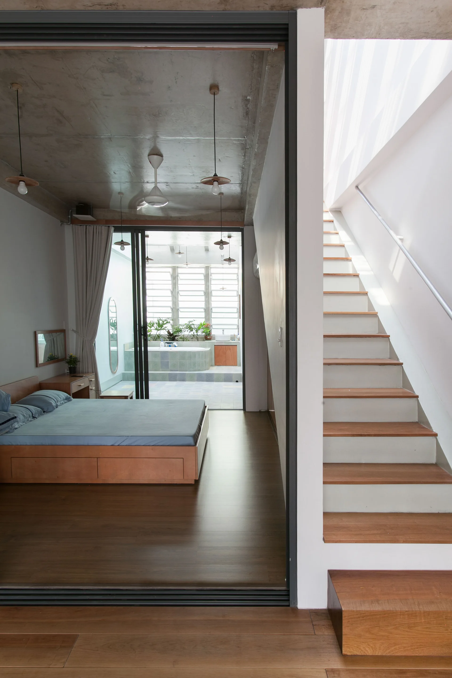 A minimalist bedroom featuring a wooden platform bed, a raw concrete ceiling, and an adjacent wooden staircase leading upstairs.