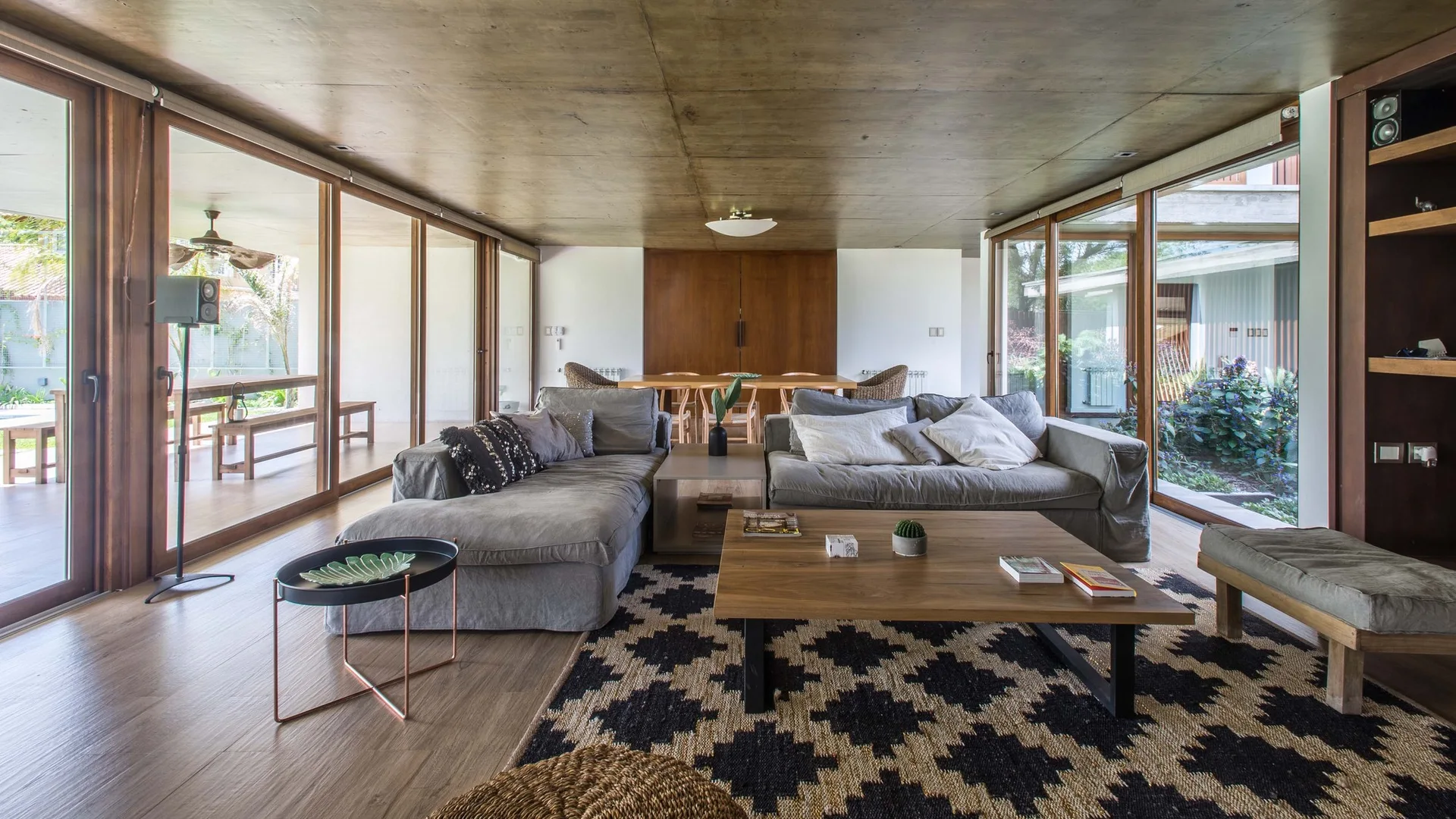 An open living room with gray sectional sofas, a wood coffee table on a patterned rug, and floor-to-ceiling glass doors showing an outdoor deck.