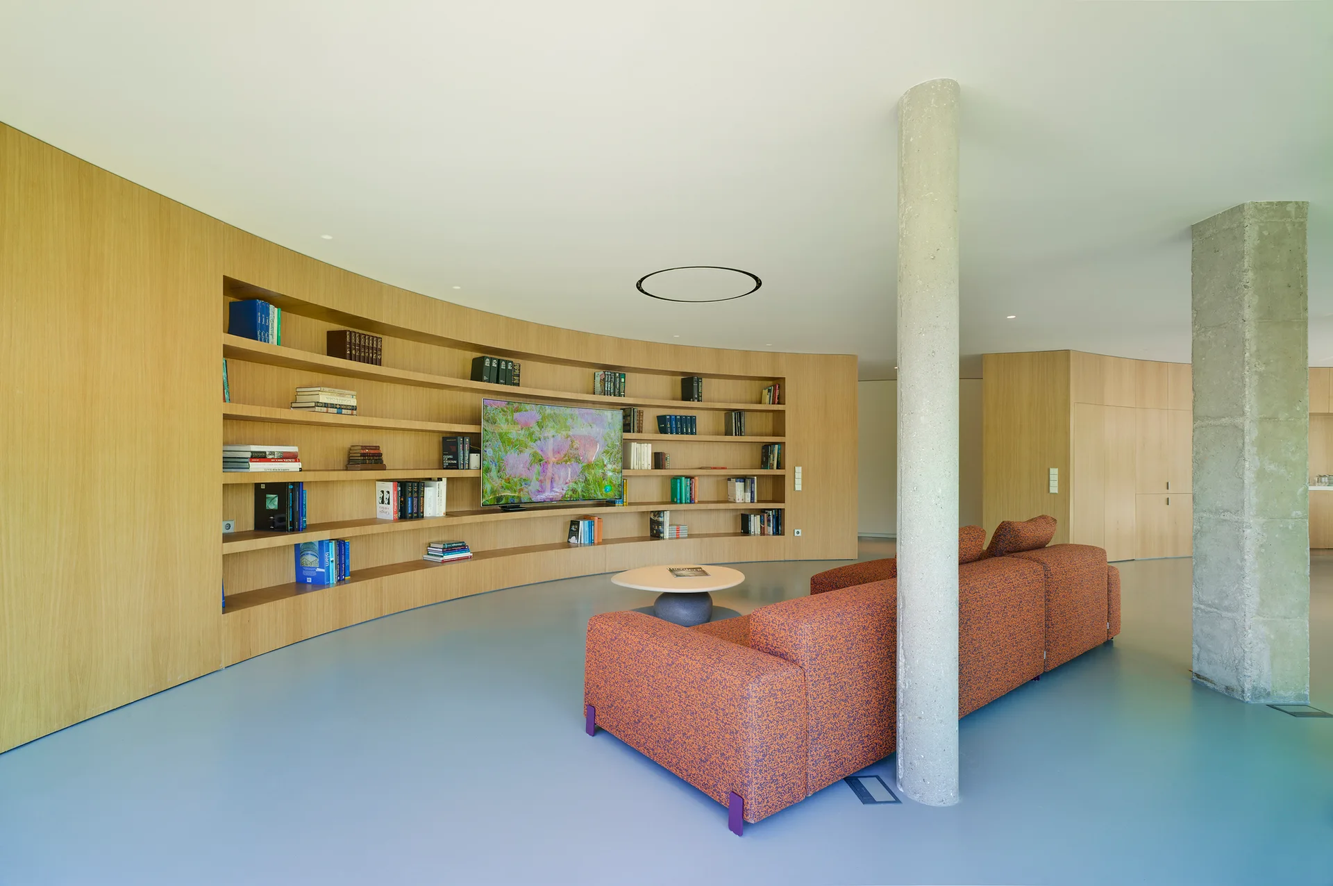 A spacious living room features a large curved wood-paneled bookshelf wall, a modern orange sofa, concrete structural columns, and smooth light blue flooring.