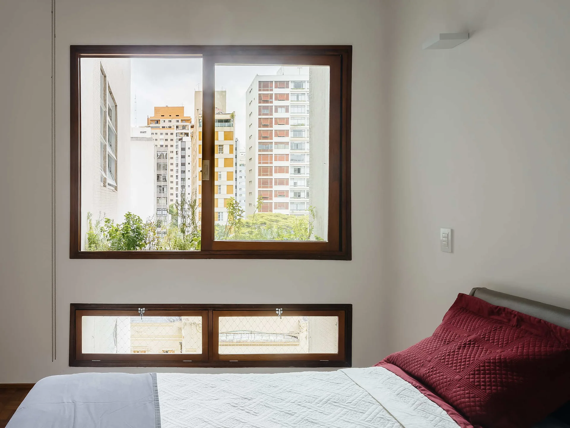 A minimalist bedroom features a bed with white and deep red bedding next to two stacked wood-framed windows overlooking city buildings.