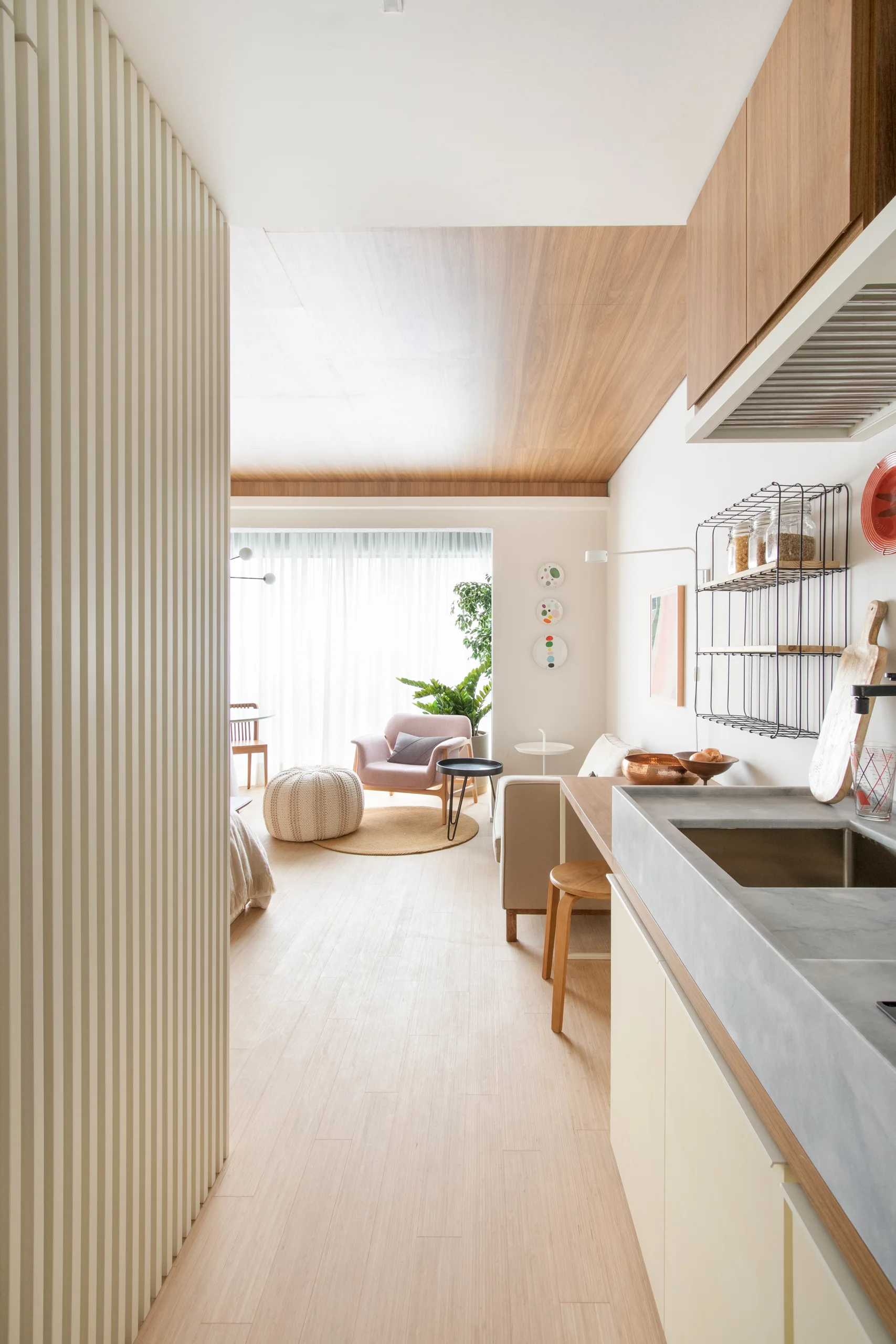 A long view of a modern apartment showing a slatted wall divider, a kitchen counter with concrete sink, and a sunlit living room with wood ceiling panels.