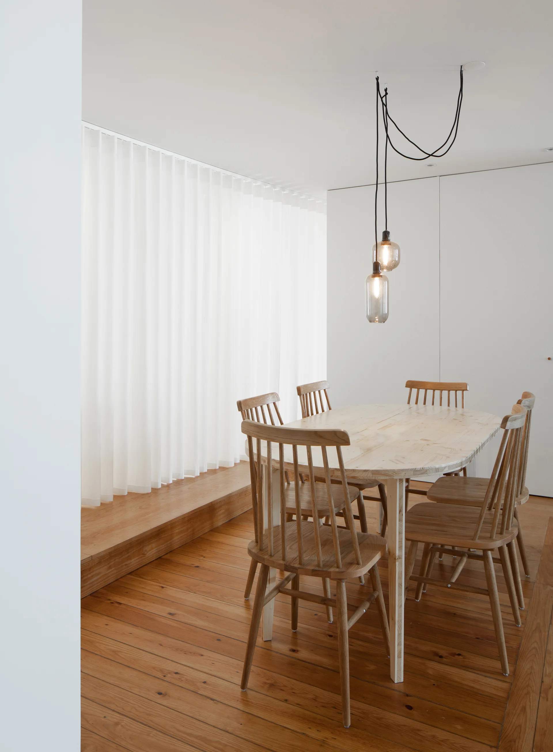 A light wood oval table and spindle chairs are positioned on wood floors next to a raised platform and sheer white curtains.