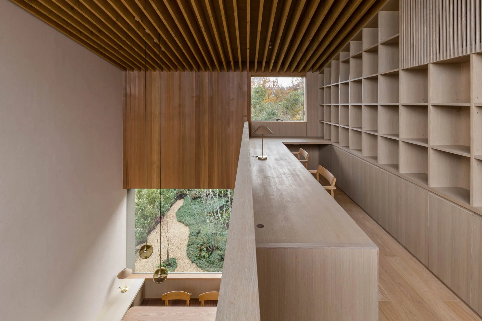 A minimalist loft library features a long light wood desk, built-in bookshelves, a slatted wooden ceiling, and a window overlooking a garden.