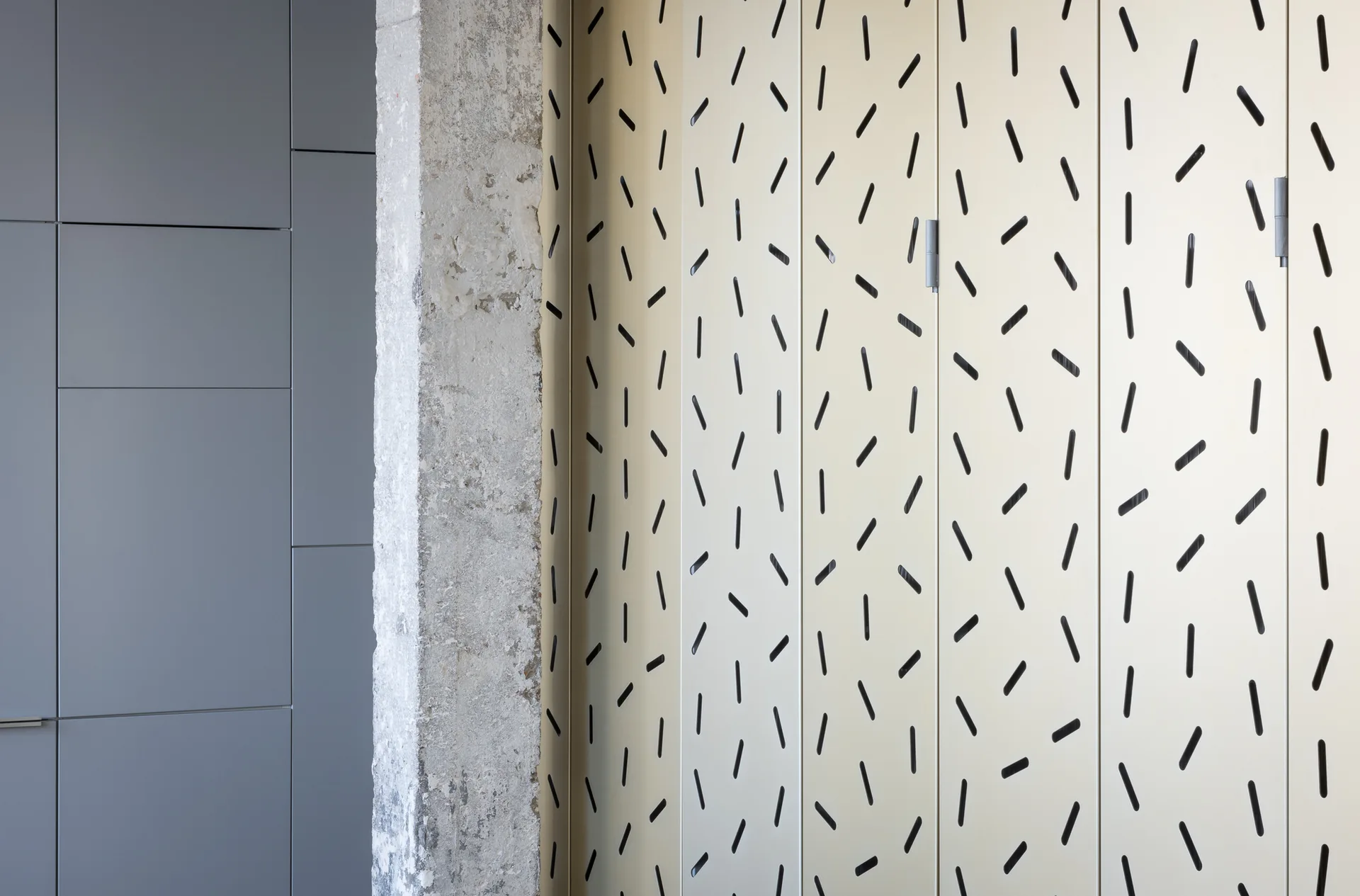 An interior wall featuring gray flat-panel cabinetry, a rough exposed concrete column, and cream metal panels with slotted perforations.