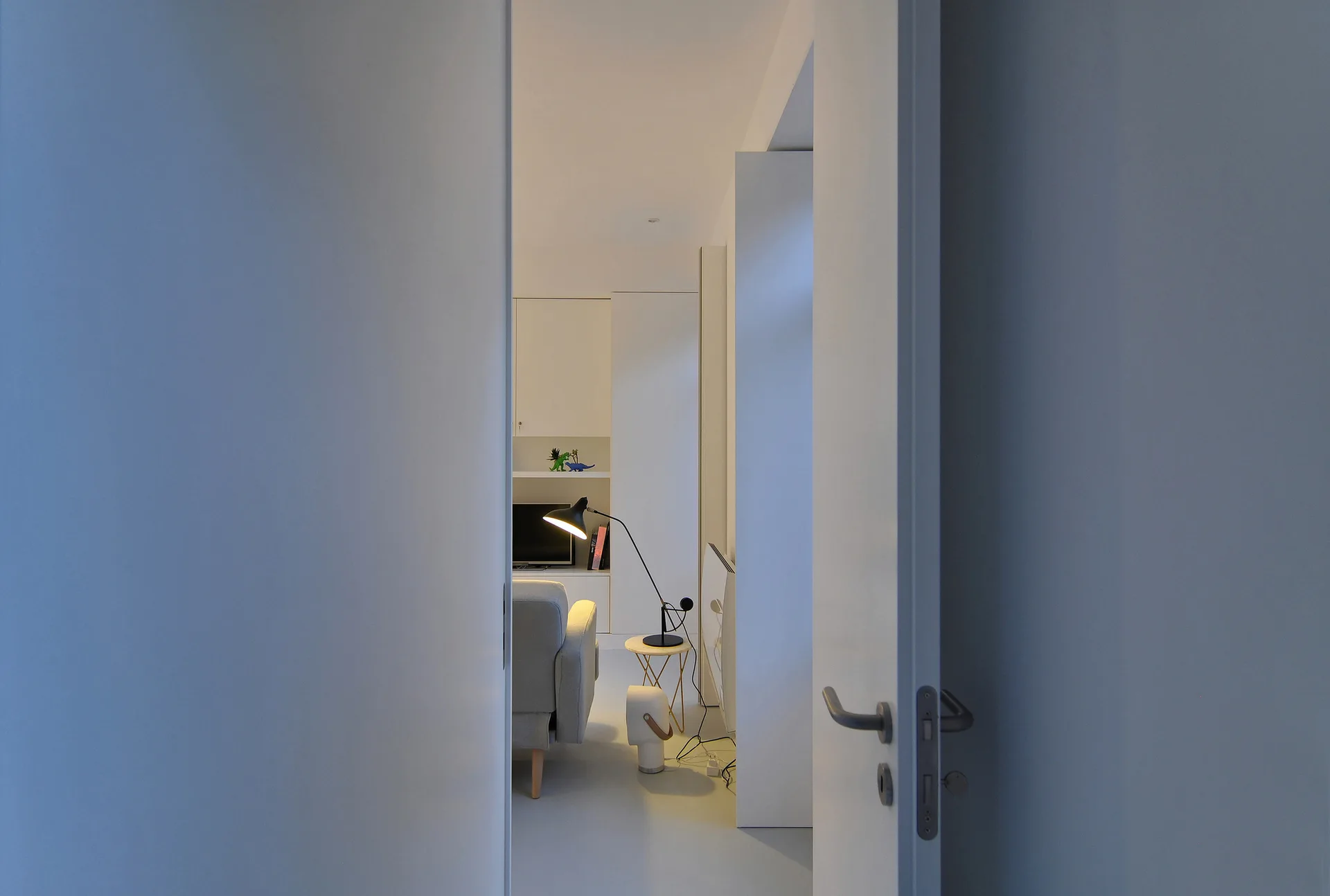 An open white door frames a view into a minimalist living room featuring a light grey sofa, a black task lamp, and white built-in storage.