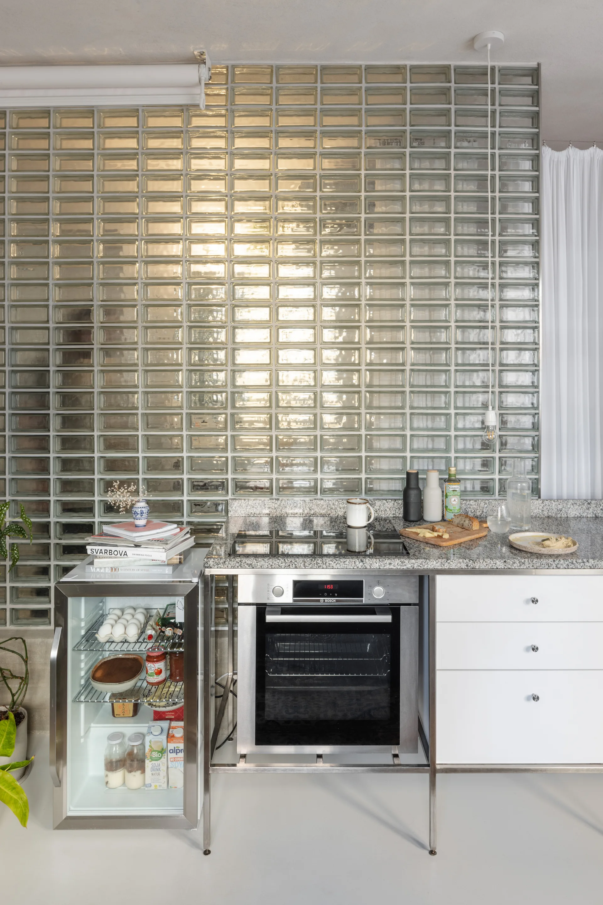 A compact kitchen features a stainless steel frame counter with a built-in oven, glass-door mini-fridge, and a floor-to-ceiling glass block wall.