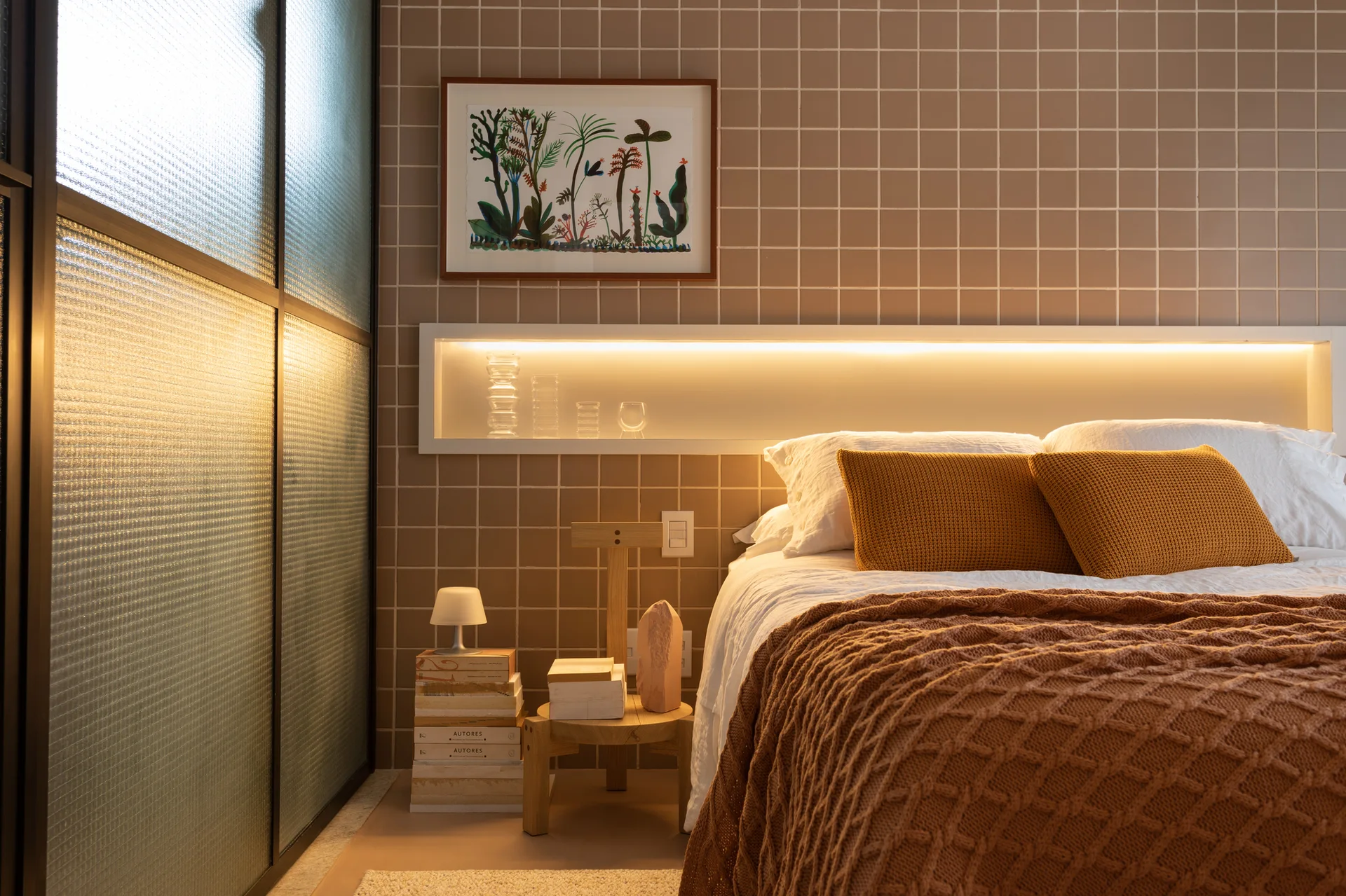 A bedroom featuring a bed with white linen and mustard pillows against a brown square-tiled wall with a recessed, illuminated shelf.