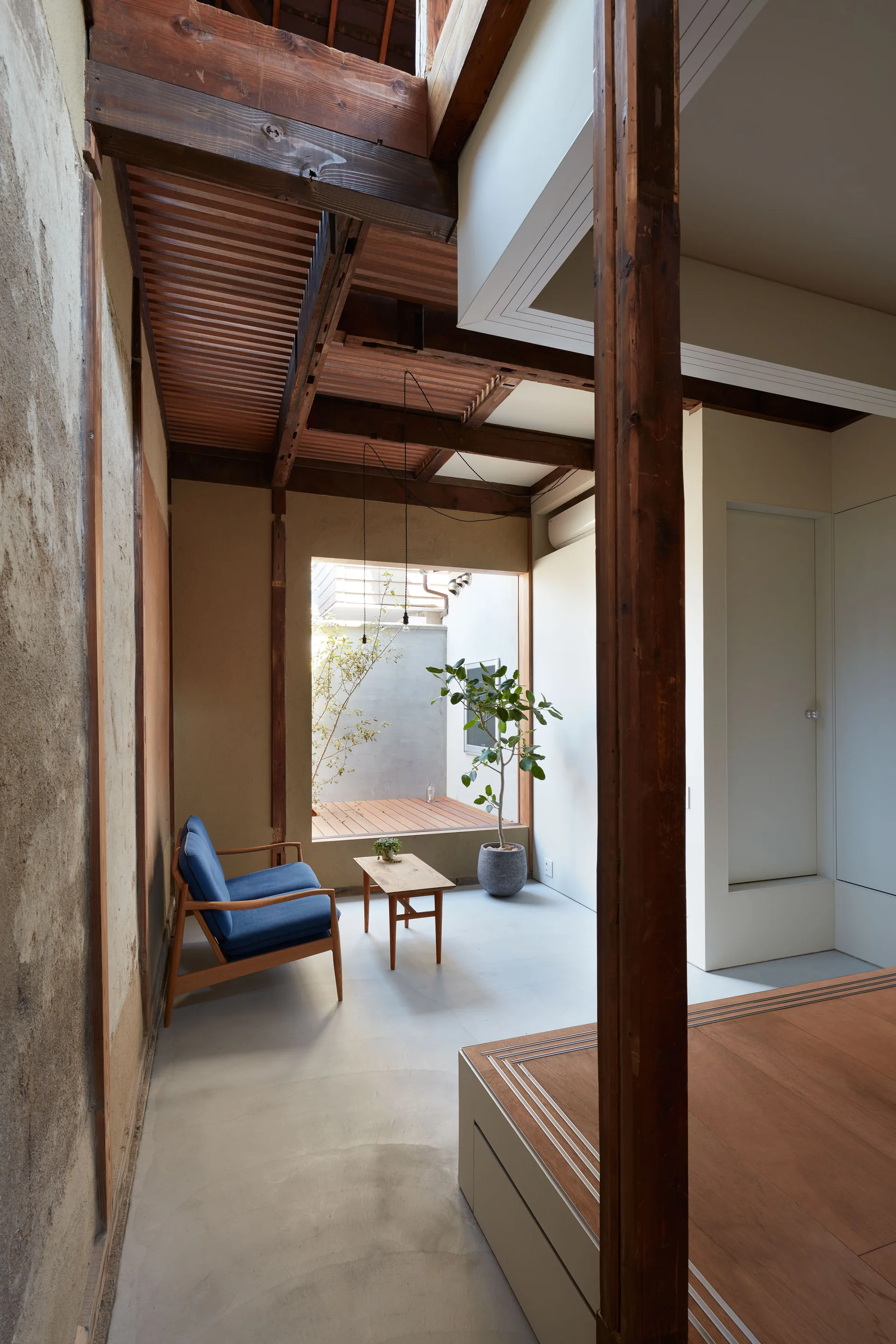 A minimalist living area with a blue armchair, a small wood coffee table, an exposed timber ceiling, and an open window leading to a courtyard.