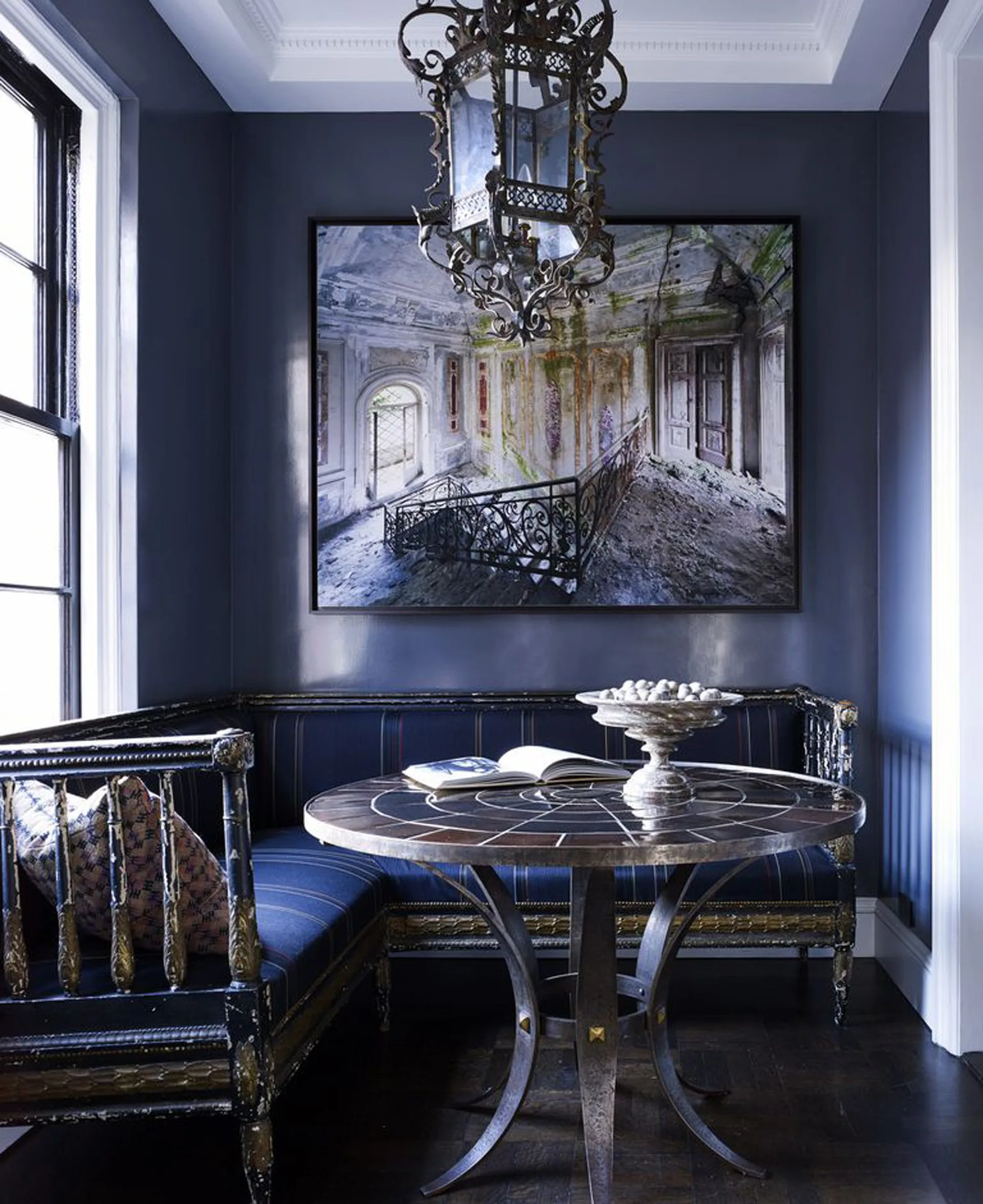 A dining corner with high-gloss navy walls, a distressed banquette, a round mosaic-topped table, and an ornate metal hanging lantern.
