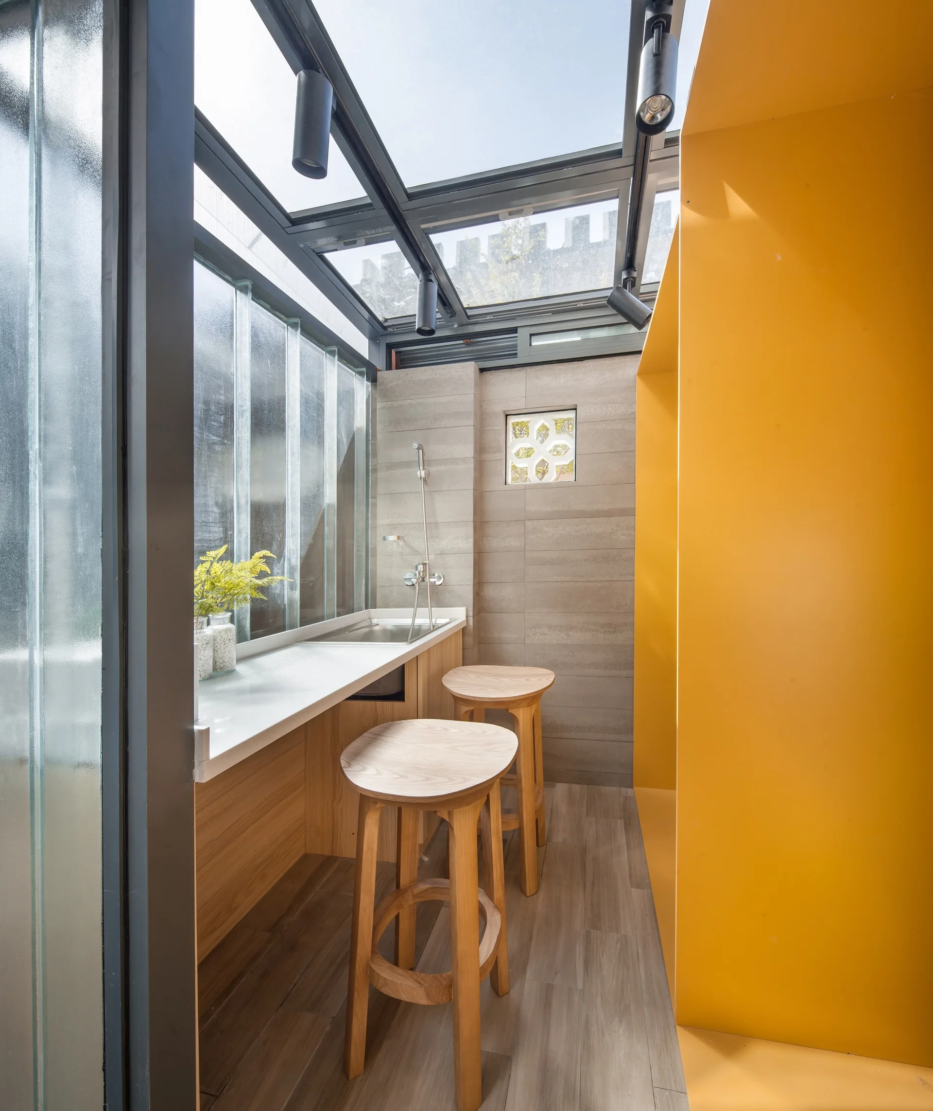 Two wooden stools stand at a white counter with a sink in a narrow room featuring a glass roof, a textured glass wall, and a yellow partition.