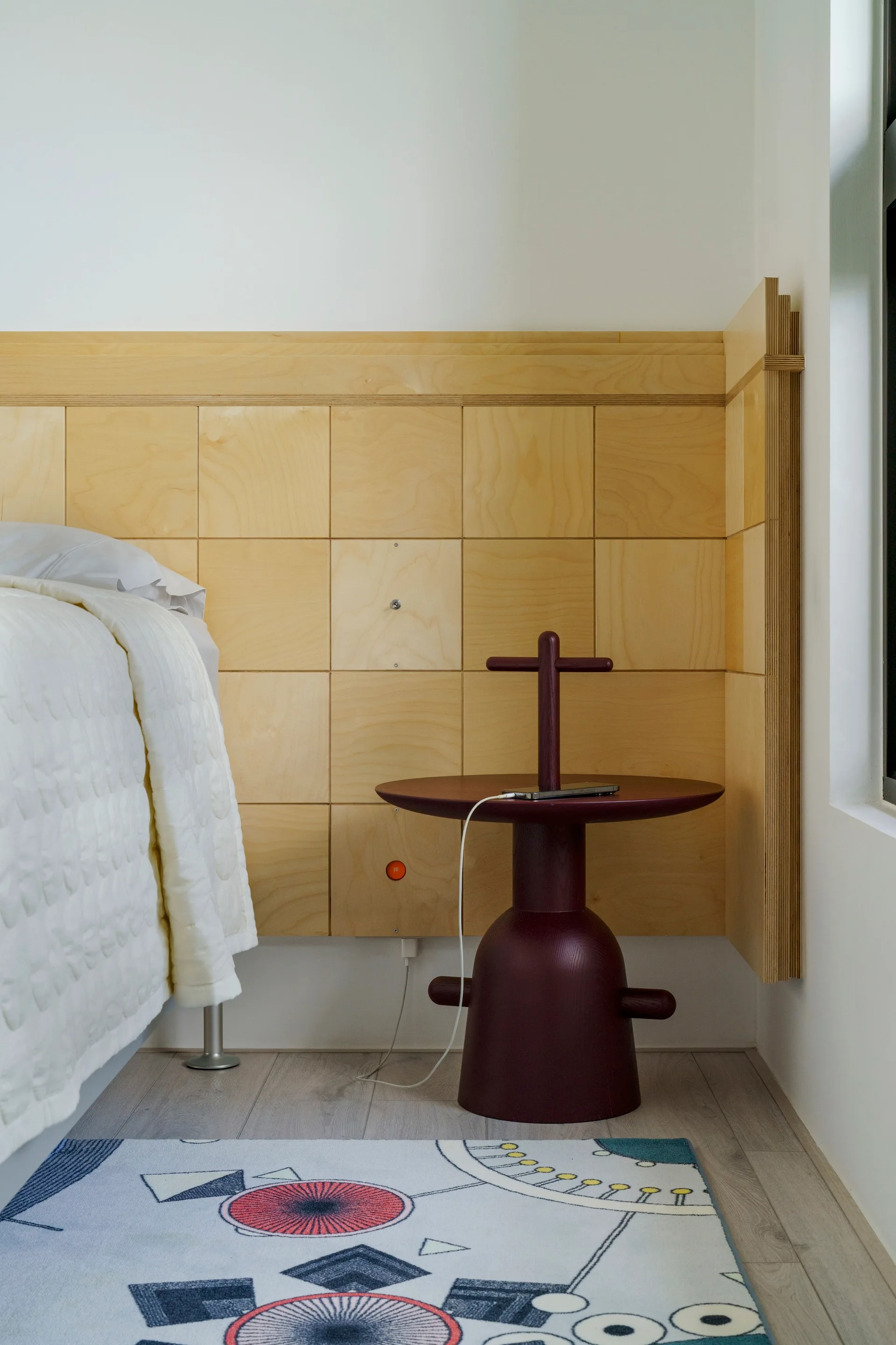 A bedroom corner features a grid-patterned light plywood headboard, a sculptural dark burgundy side table, and a patterned area rug next to white bedding.