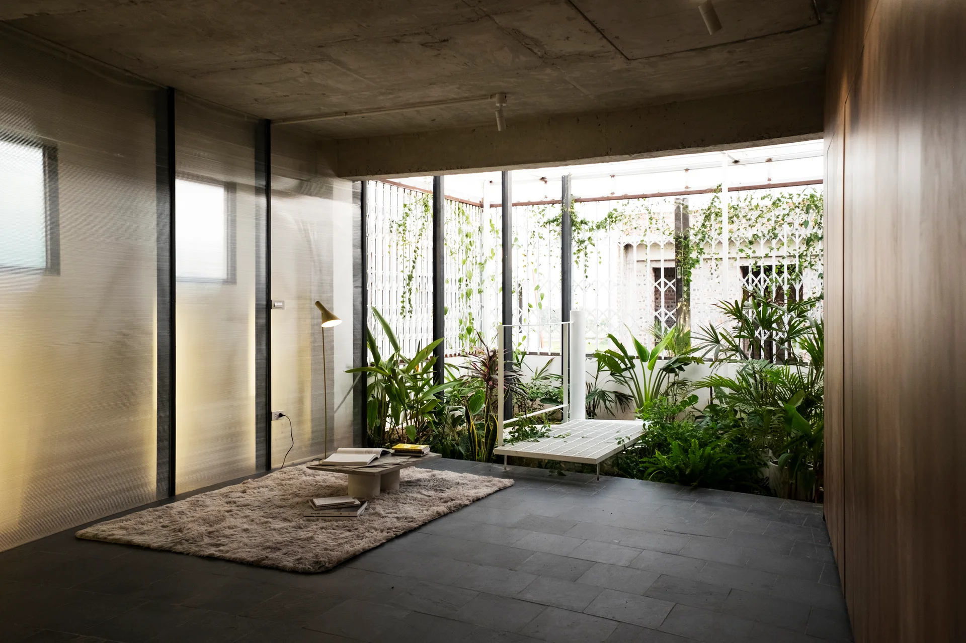 A minimalist living space features a fluted translucent wall, a raw concrete ceiling, and a lush indoor courtyard atrium filled with green plants.