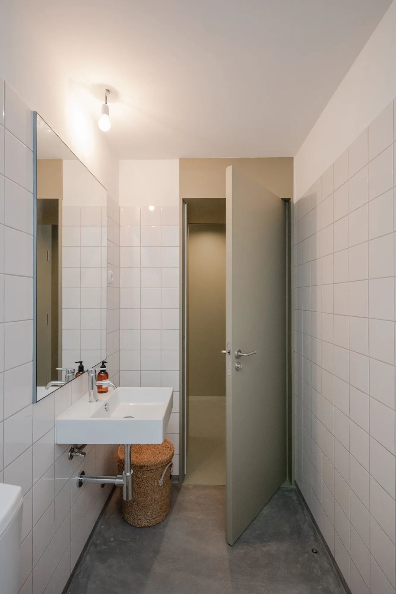 A minimalist bathroom with glossy white square grid tiles on the walls, a wall-mounted sink, and an open sage green door.