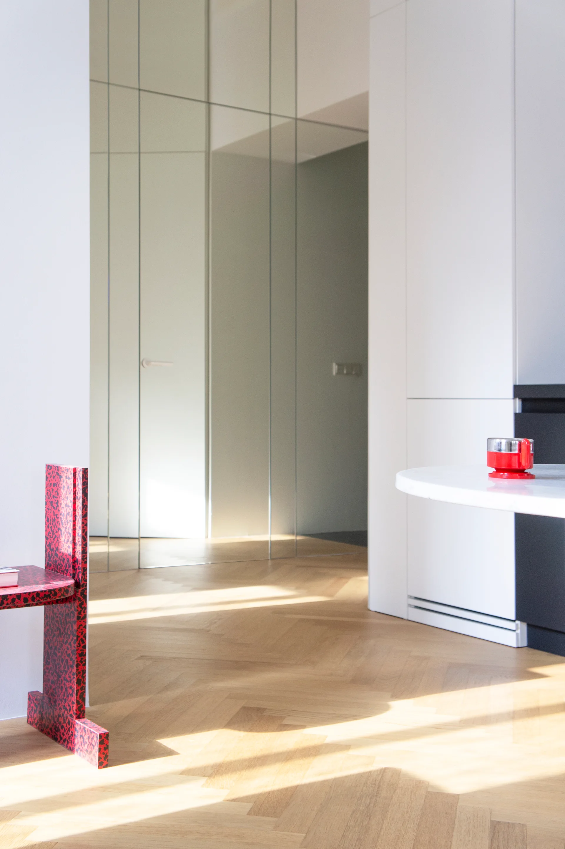 A modern interior entryway featuring a graphic red speckled chair, herringbone hardwood floors, and a floor-to-ceiling mirrored wall panel.