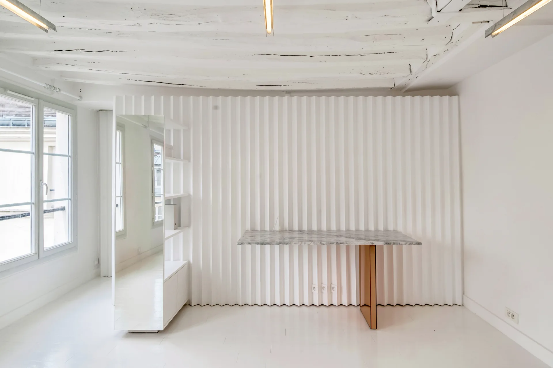 A minimalist white office featuring a marble-topped desk, a vertical mirror, and a textured corrugated feature wall under exposed ceiling beams.
