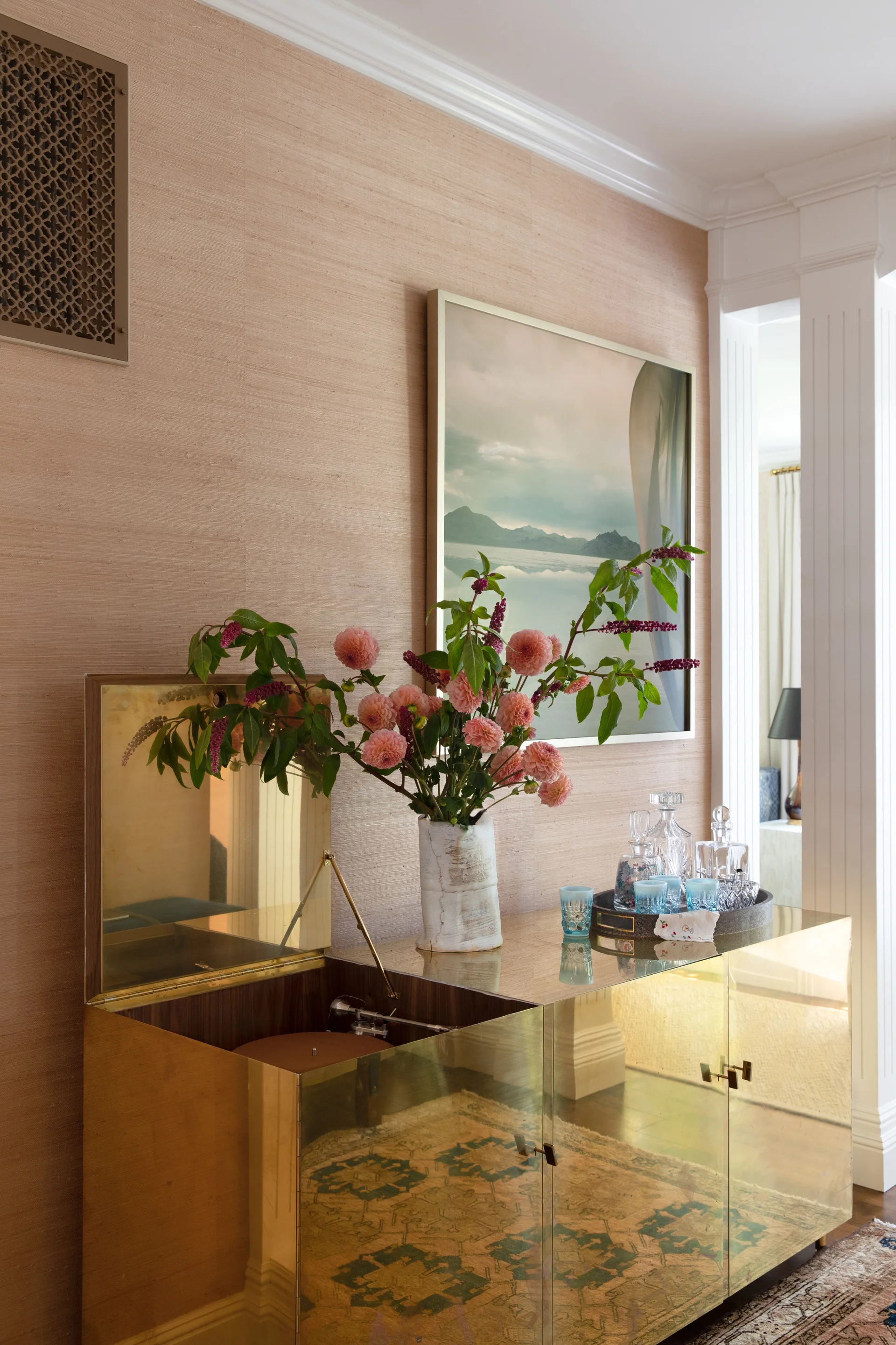 A metallic brass bar cabinet with an open top showing a turntable sits against a beige grasscloth wall beneath a framed landscape artwork.