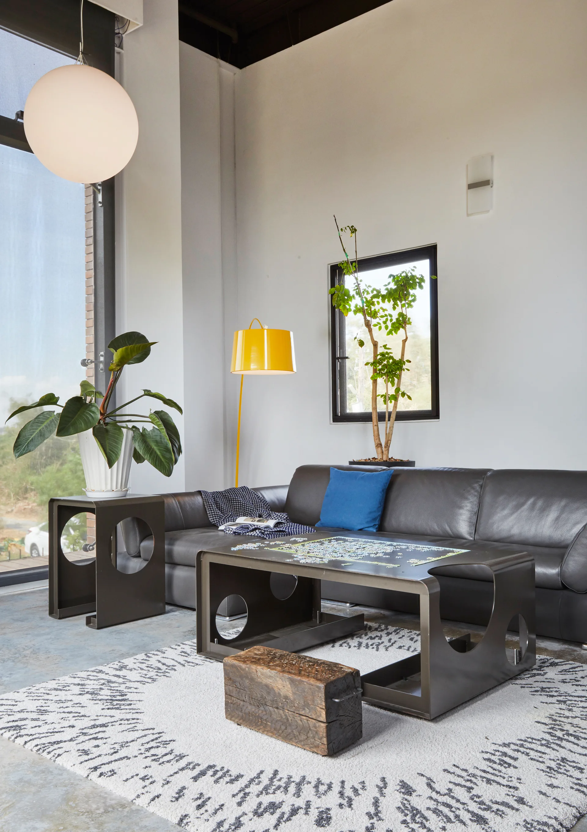 A modern living room features a dark leather sofa, geometric metal tables, a patterned white area rug, and a bright yellow floor lamp.