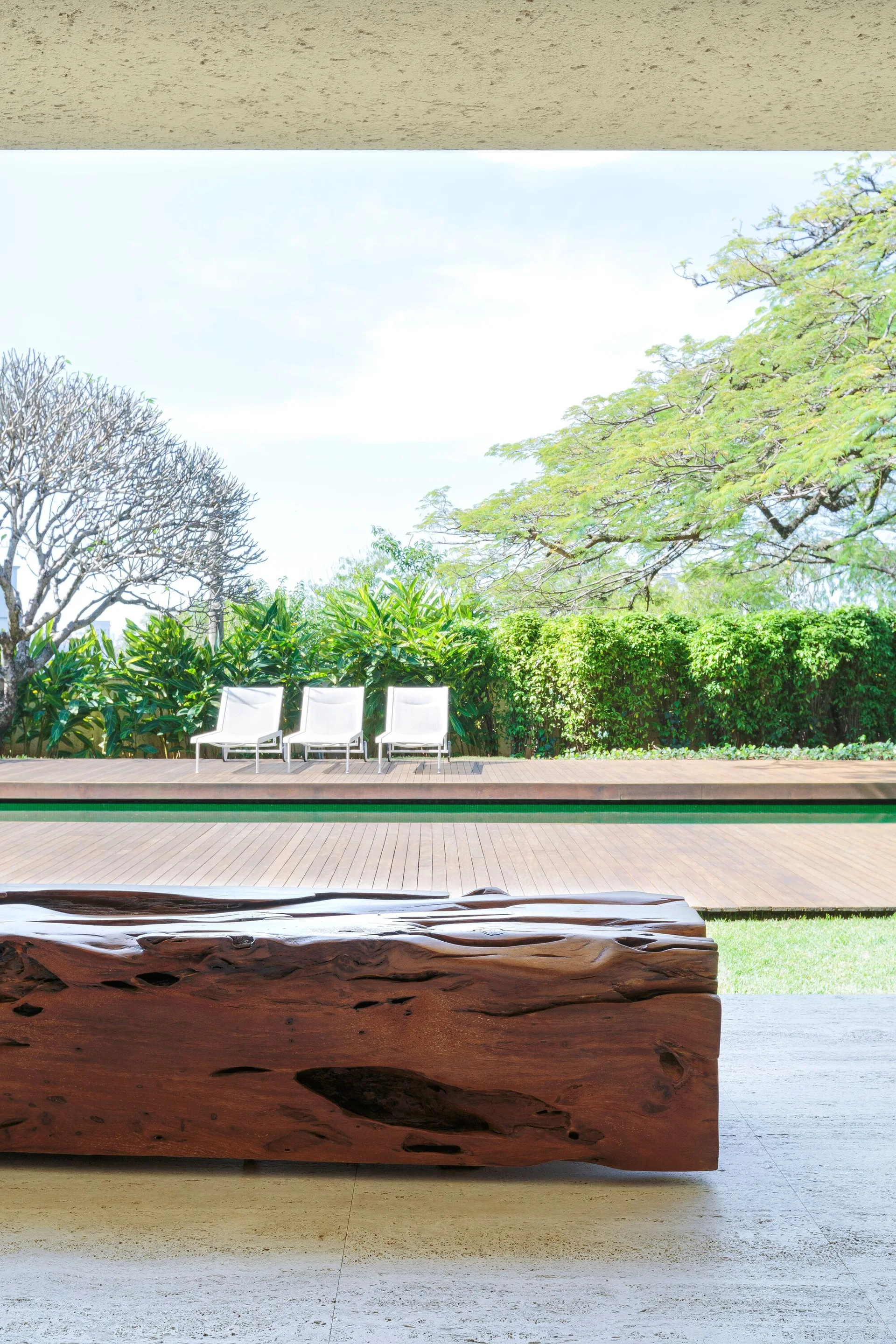 An outdoor patio features a large raw wood bench in the foreground looking out toward a swimming pool with lounge chairs.