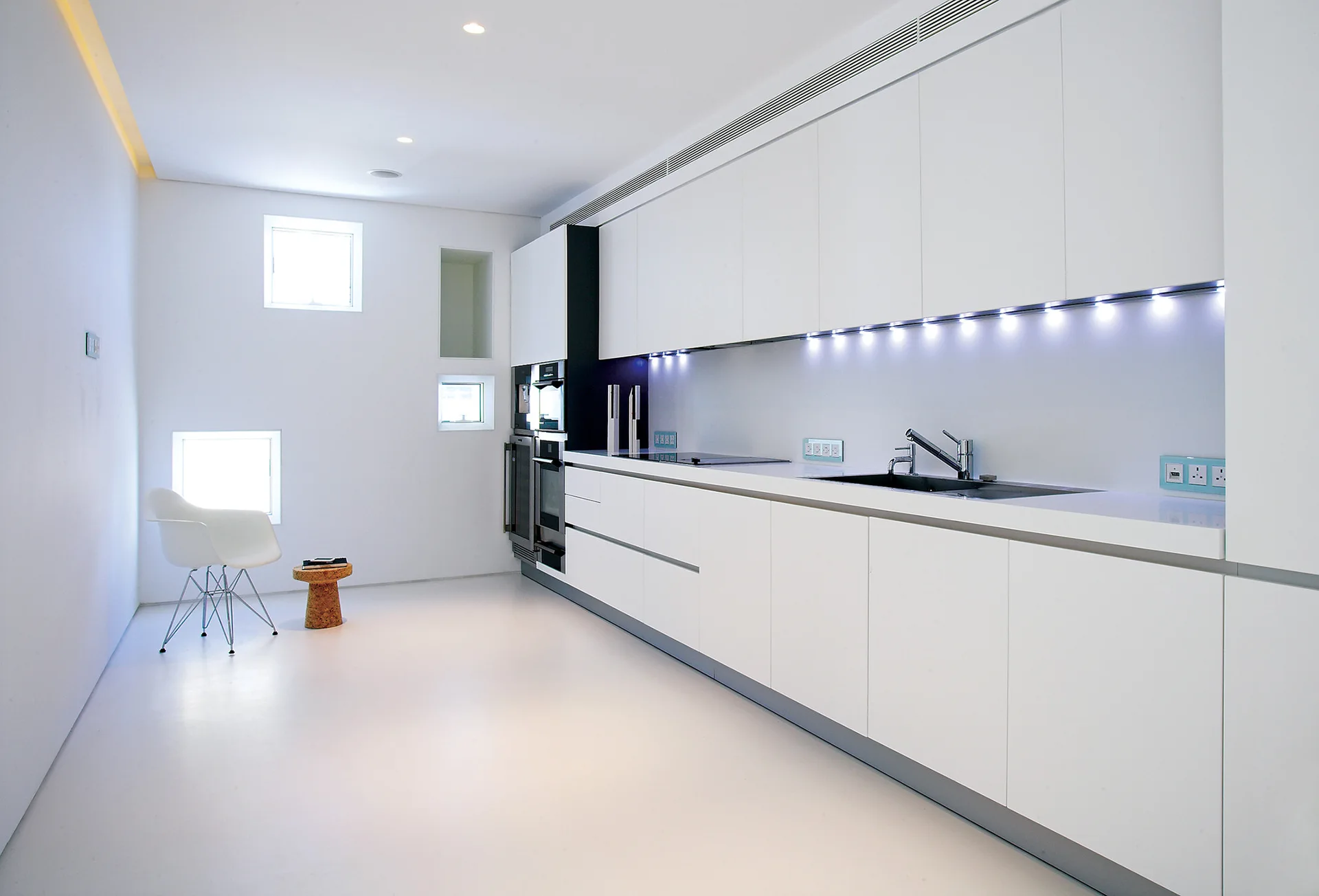 A modern white kitchen features sleek handleless cabinets illuminated by under-cabinet LED lighting, built-in appliances, and small square windows.