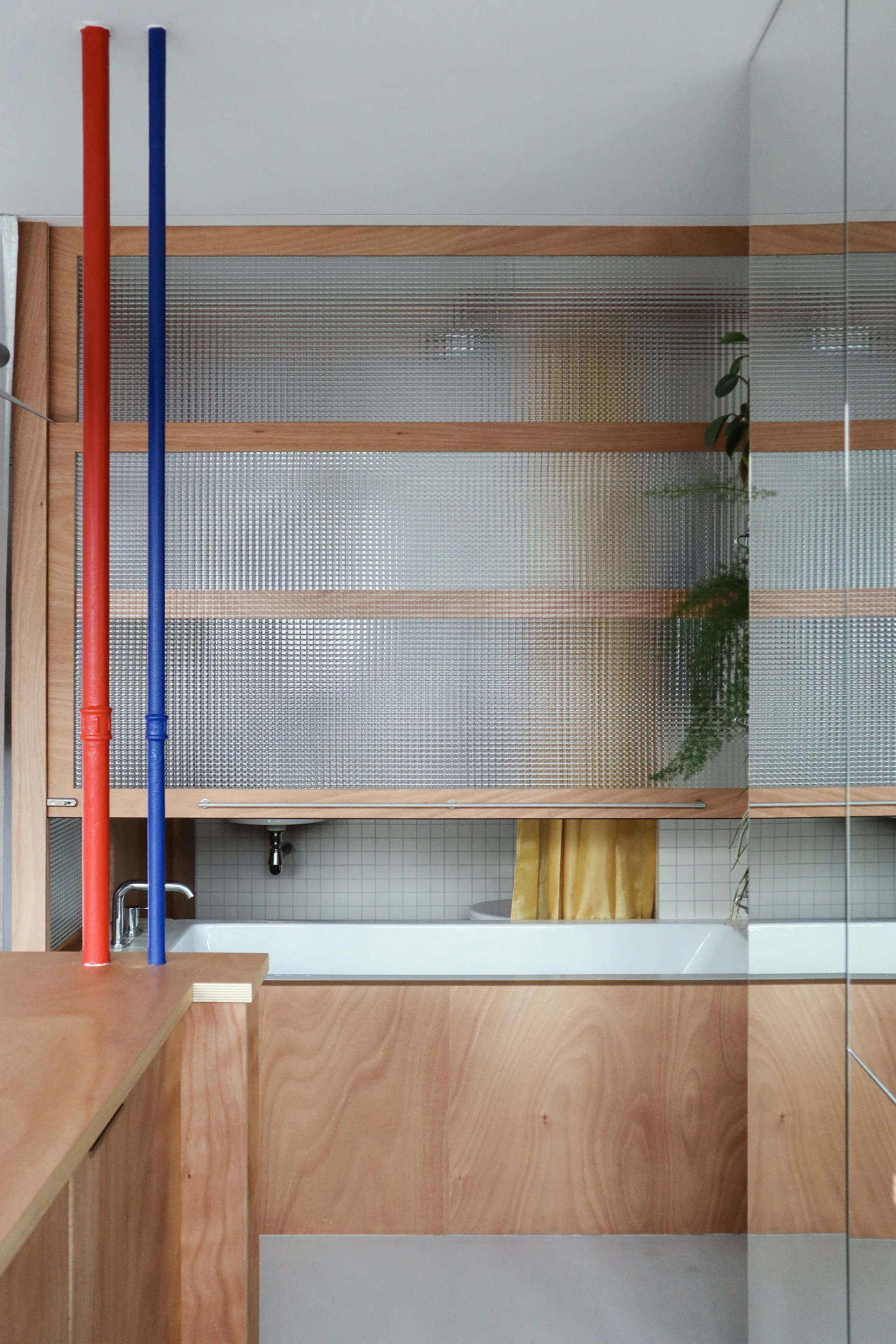 A modern bathroom featuring a plywood-wrapped bathtub, a textured glass partition screen, and prominent red and blue vertical plumbing pipes.