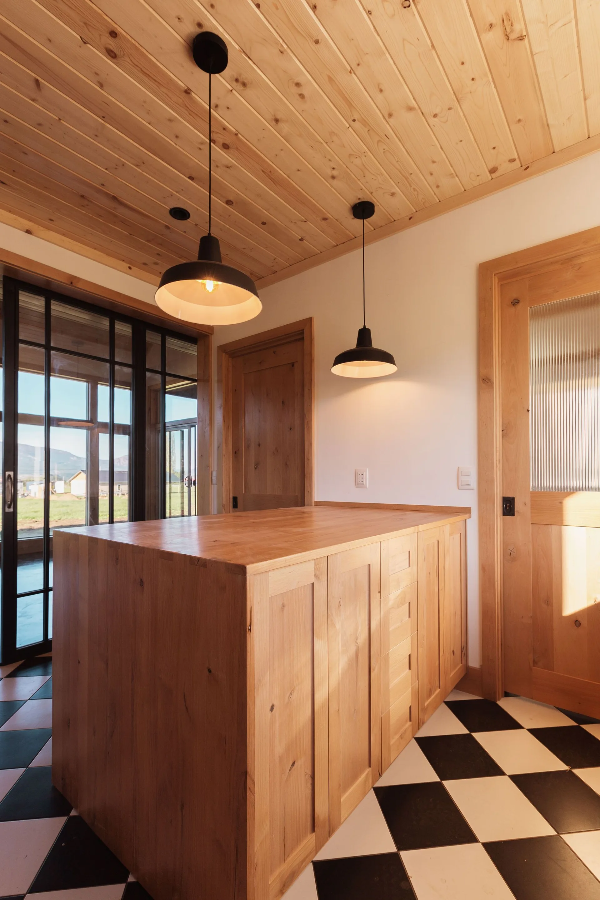 A light wood kitchen island sits on a black and white checkered floor beneath a matching wood plank ceiling and black pendant lights.