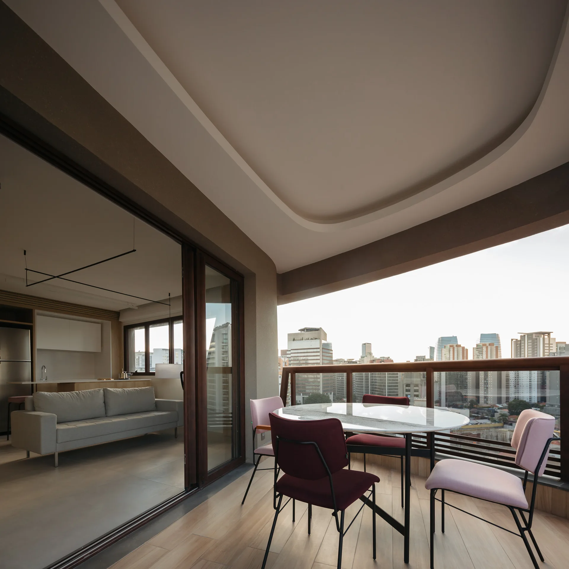 A modern apartment balcony features a round dining table with pink and burgundy chairs next to sliding glass doors showing an indoor living room.
