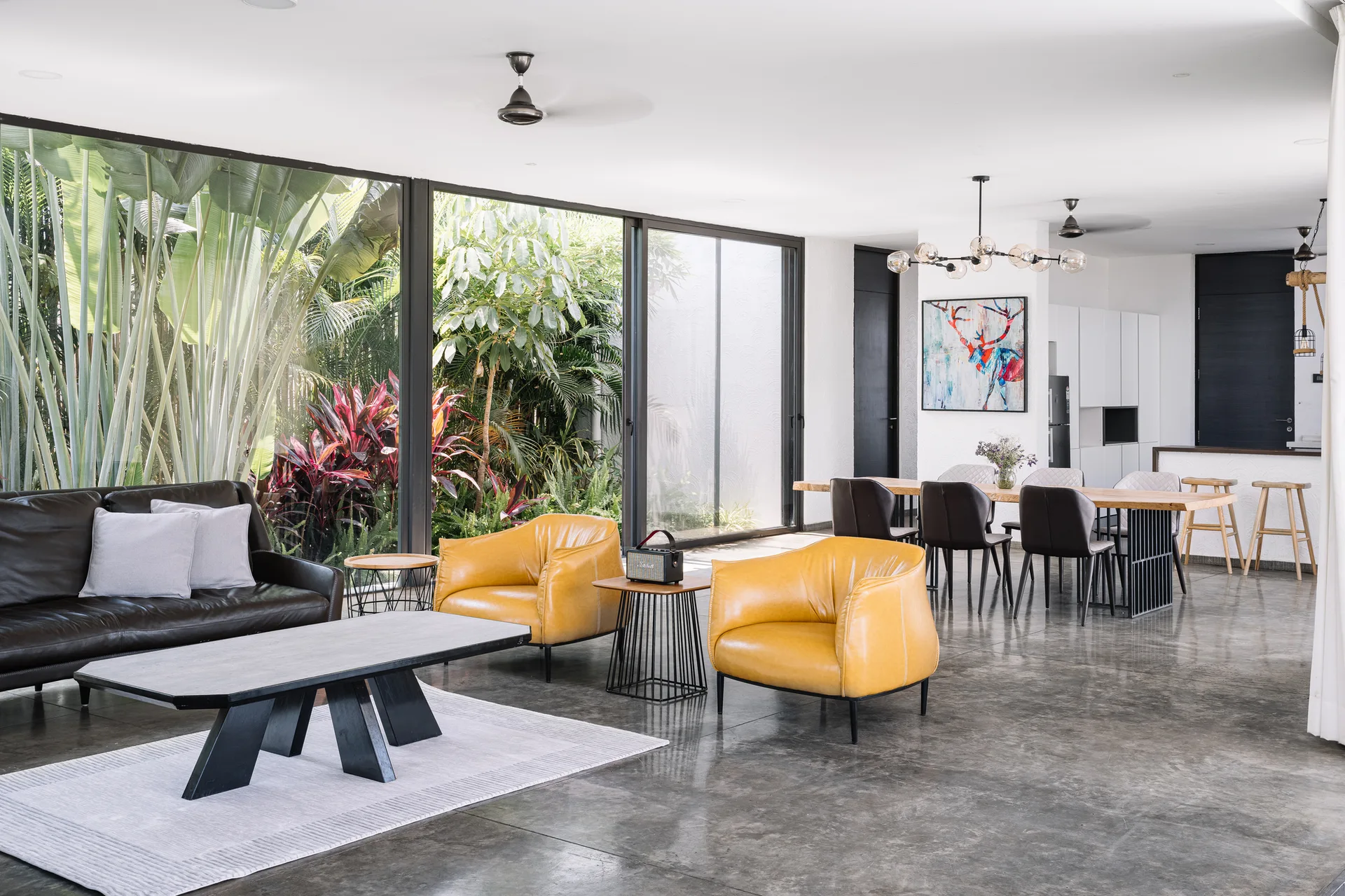 A modern open-concept living room features yellow leather armchairs, a dark sofa, polished concrete floors, and large glass windows showing a lush garden.