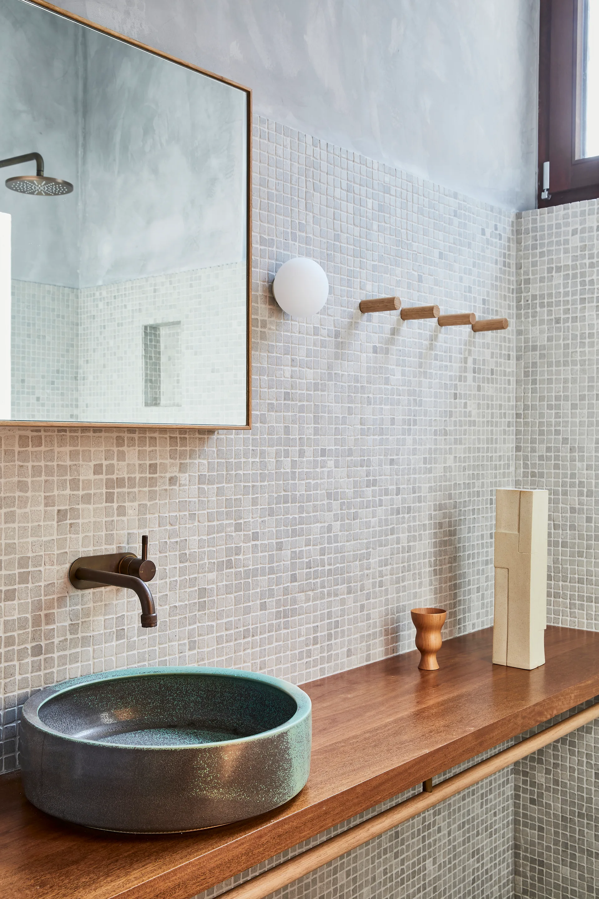 A minimalist bathroom vanity features a wood counter, a teal vessel sink, a wall-mounted faucet, and a wall covered in small gray square mosaic tiles.