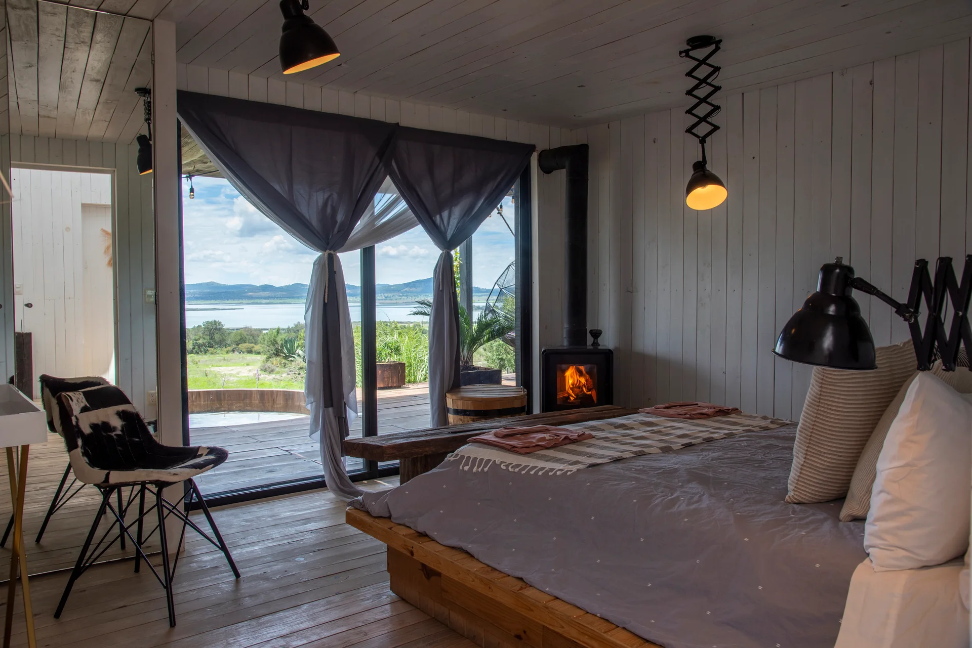 A rustic bedroom features a platform bed, vertical white wood paneled walls, a black wood stove, and sliding glass doors looking out to a scenic lake.