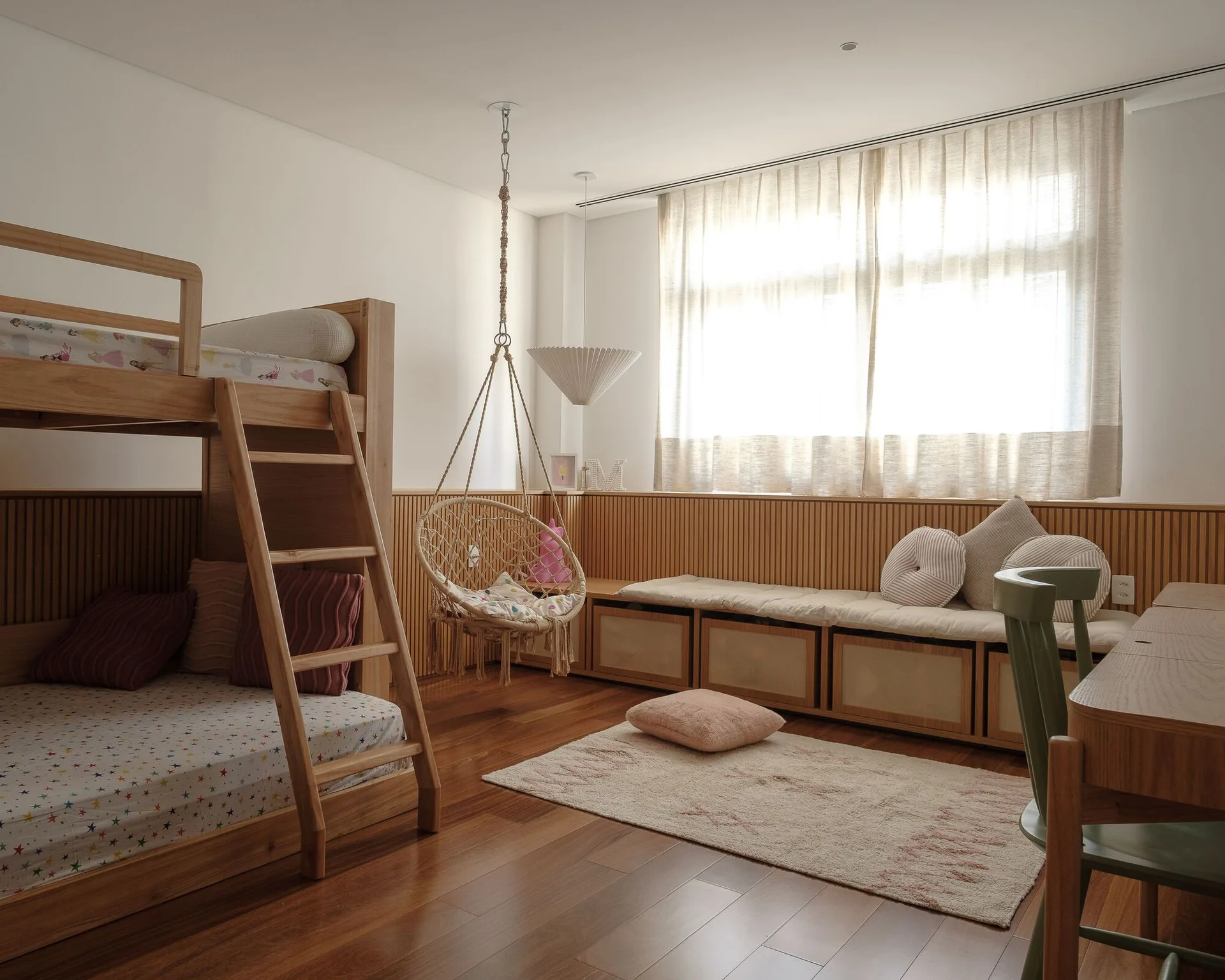 A children's bedroom featuring a wooden bunk bed, a hanging rope chair, and a long built-in storage bench against a slatted wood paneled wall.