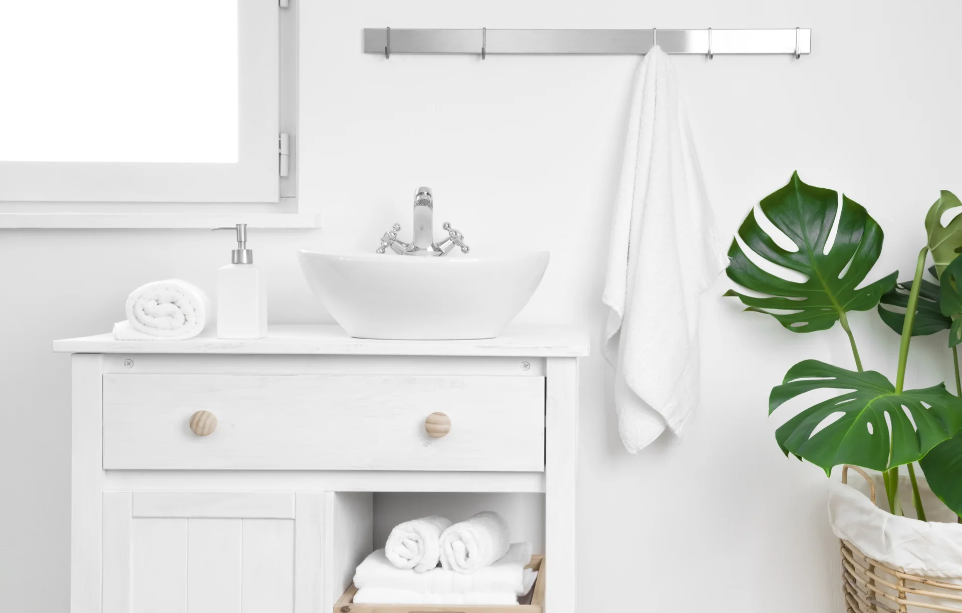 A clean white bathroom vanity features a ceramic vessel sink and rolled white towels, situated next to a potted monstera plant.