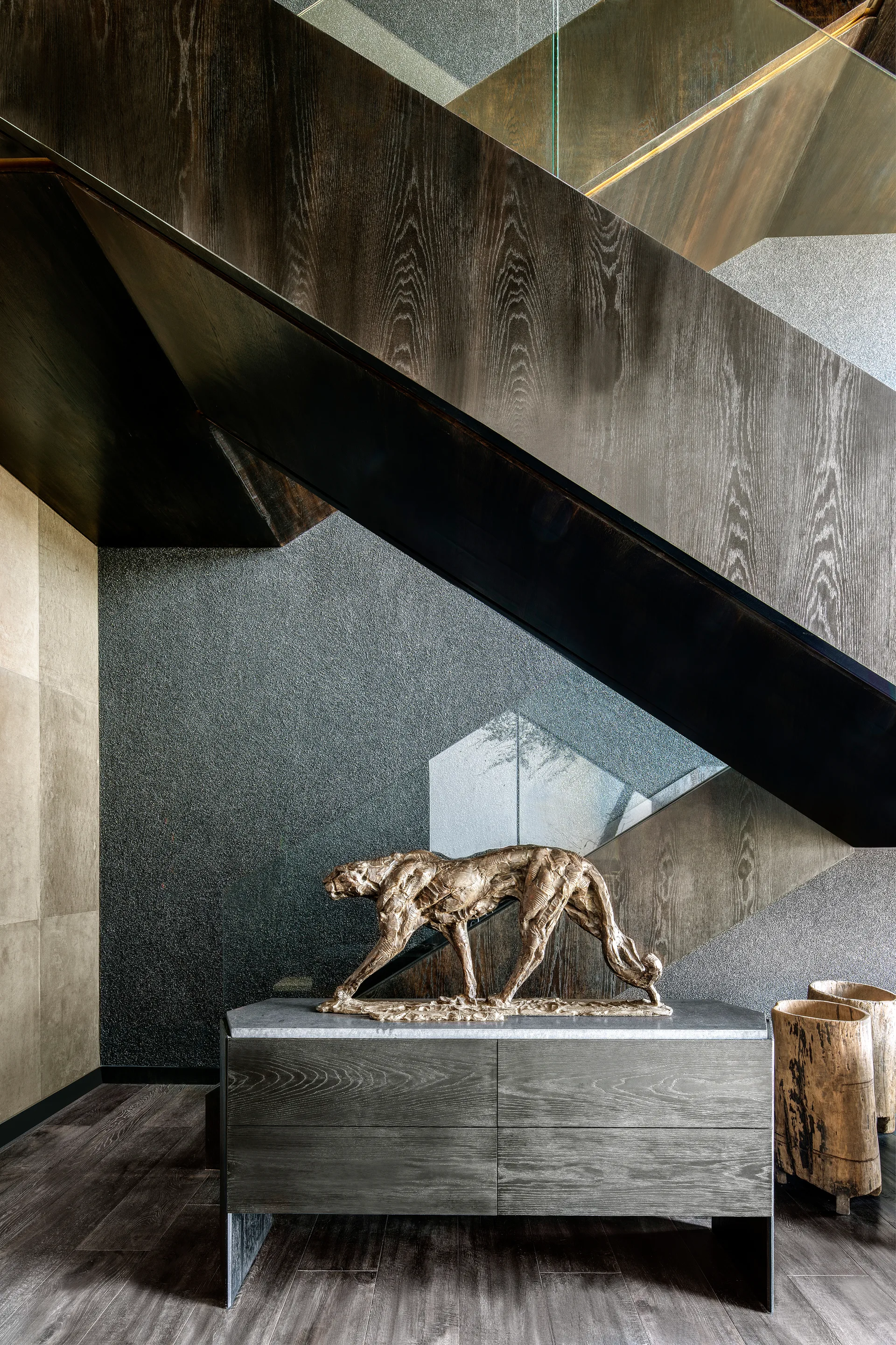A dark wood staircase with a glass railing ascends next to a stone-topped wood console displaying a bronze animal sculpture.