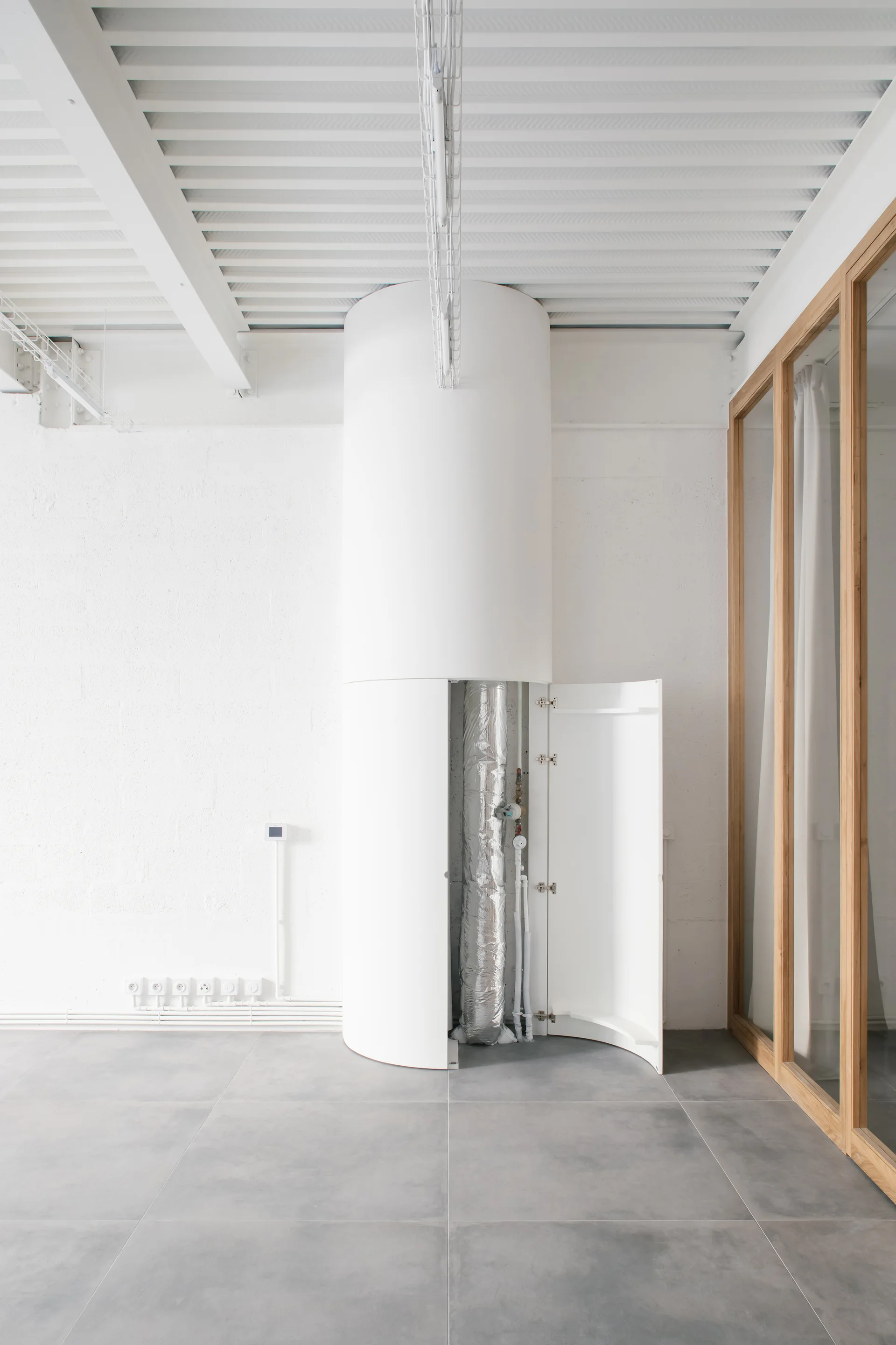 An open cylindrical white utility cabinet revealing insulated pipes next to a wood-framed glass partition on a grey tiled floor.